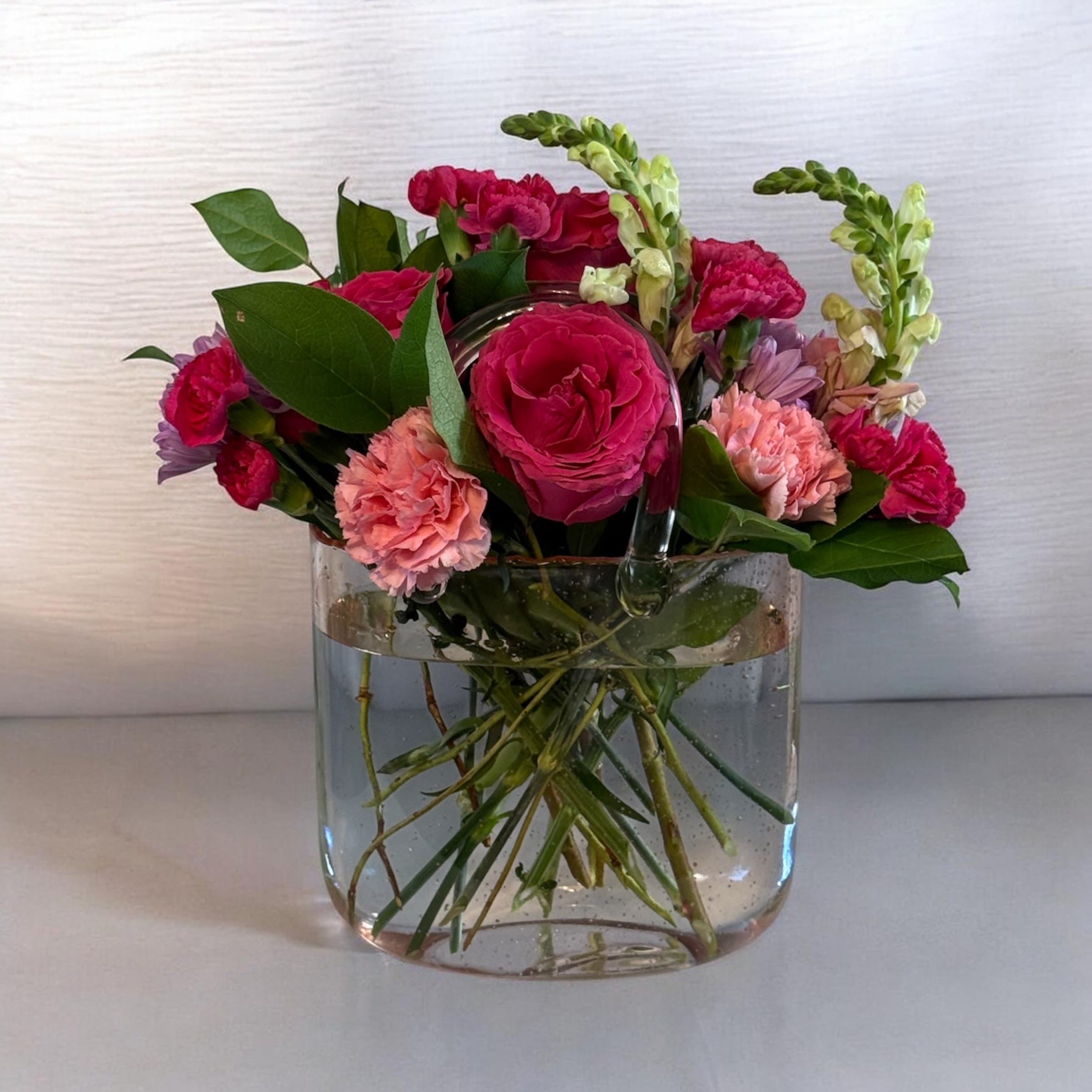 Petal Perfection's "Let my get my Purse" in Pink - Bright pink roses and coral carnations form a joyful, compact cluster in this playful floral arrangement-perfect as a Mother's Day gift, birthday bouquet, a cheerful thank-you gift, or a springtime surprise. Designed as an everyday celebration piece, "Let me get my Purse" in Pink presents a vivid, feminine palette of pink, red, peach, white, purple, and glossy green foliage that immediately reads as bright and affectionate to the person you're thinking of.  The design pairs rounded blooms-roses, carnations, and small daisy-like flowers-with upright snapdragon spikes for height and movement, creating a lively, slightly asymmetrical silhouette. The handbag-shaped pink glass vase is a whimsical, gift-ready look; arranged densely so the bouquet reads full and abundant.   This arrangement is ideal for moms, friends, coworkers, or anyone who should feel seen and celebrated-its bright colors and fresh textures say "you made my day" without words. Standard care: keep water fresh and trim stems every few days to prolong bloom life; select your preferred vase option at checkout for immediate gifting-ready presentation.