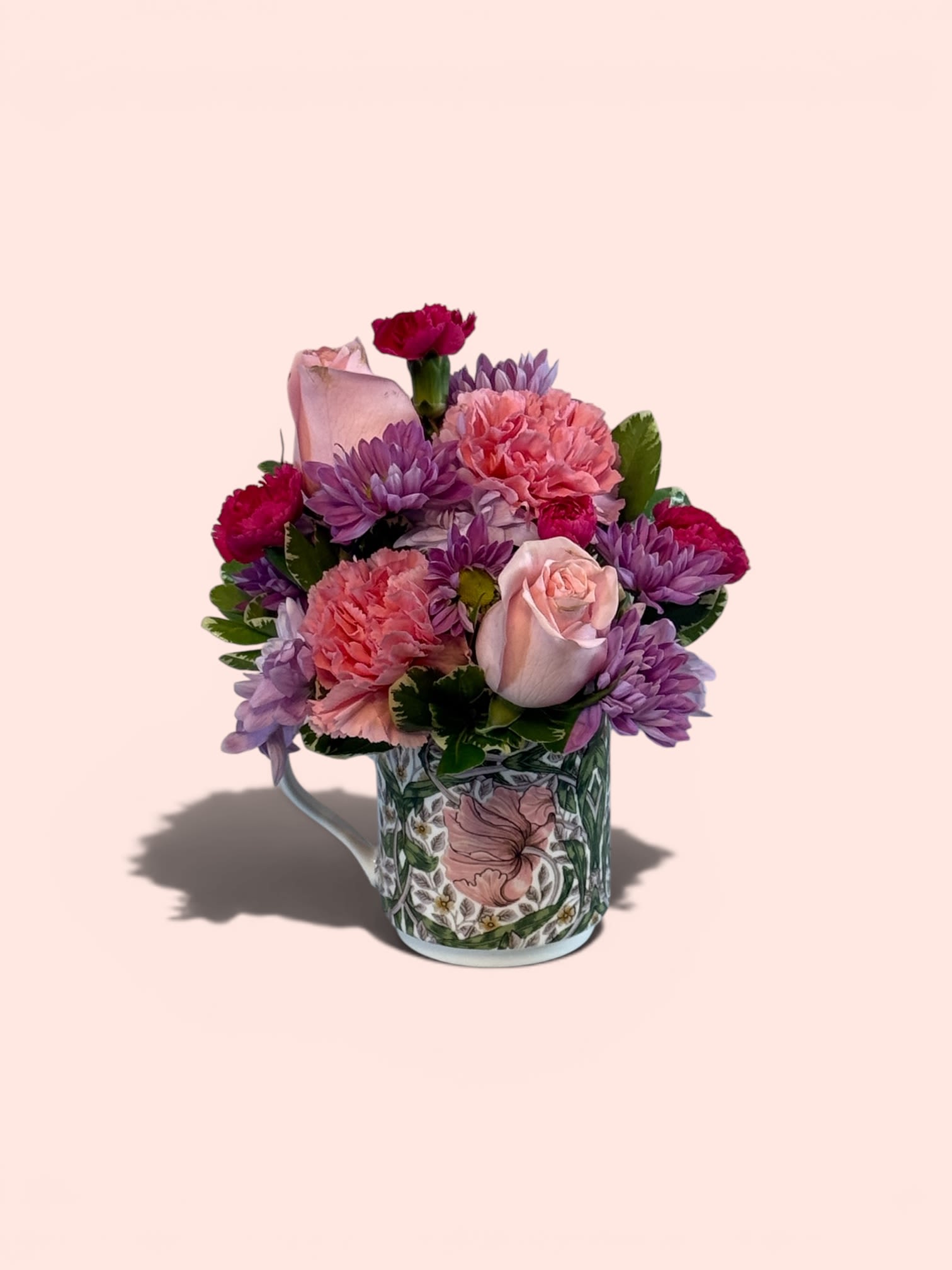 Mom's New Favorite Mug a Petal Perfection Best Seller - A compact cluster of soft pink carnations, pale pink roses, and vivid purple chrysanthemums fills a floral-patterned ceramic mug-an ideal Mother' Day, birthday bouquet, thank-you gift, or "just because" arrangement for Mom. The pink, purple, white, and green palette reads cheerful and gently romantic, the kind of everyday gift that makes the person you care about feel noticed and cherished.  Flowers are arranged tight and rounded for a full, tabletop-ready silhouette; the decorative ceramic mug adds a casual, giftable touch. Small-scale and easy to display, this arrangement suits bedside tables, kitchen counters, or office desks-no special care beyond fresh water and a cool spot keeps the blooms looking their best.