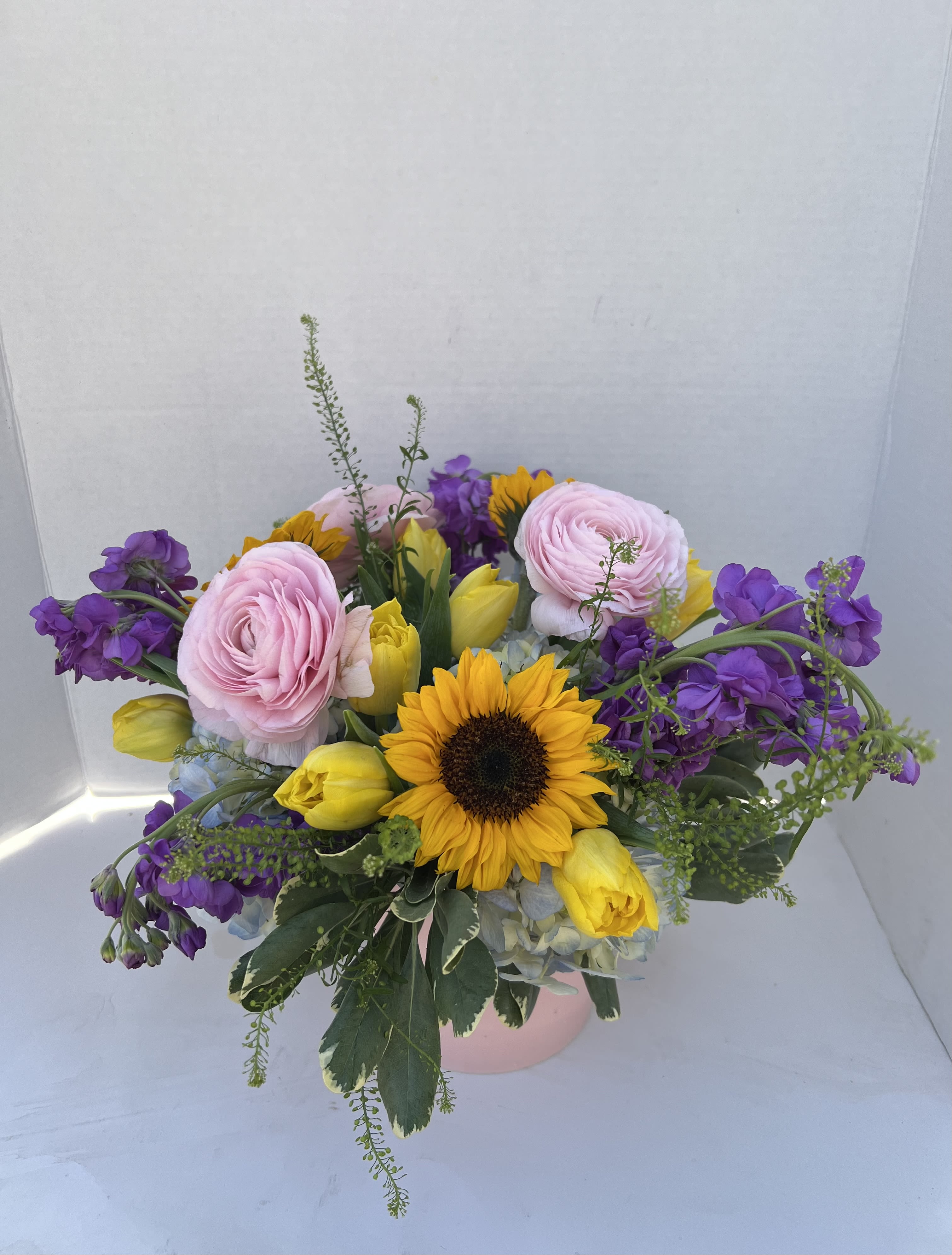 Sunny Day - Whether it’s brightening a kitchen table, celebrating a special moment, or sending a little happiness just because, Sunny Day delivers warmth, color, and joy in every petal. Sunny sunflowers, lush blue hydrangea, yellow tulips and delicately layered ranunculus blooms weave throughout the design in a pink ceramic pot.