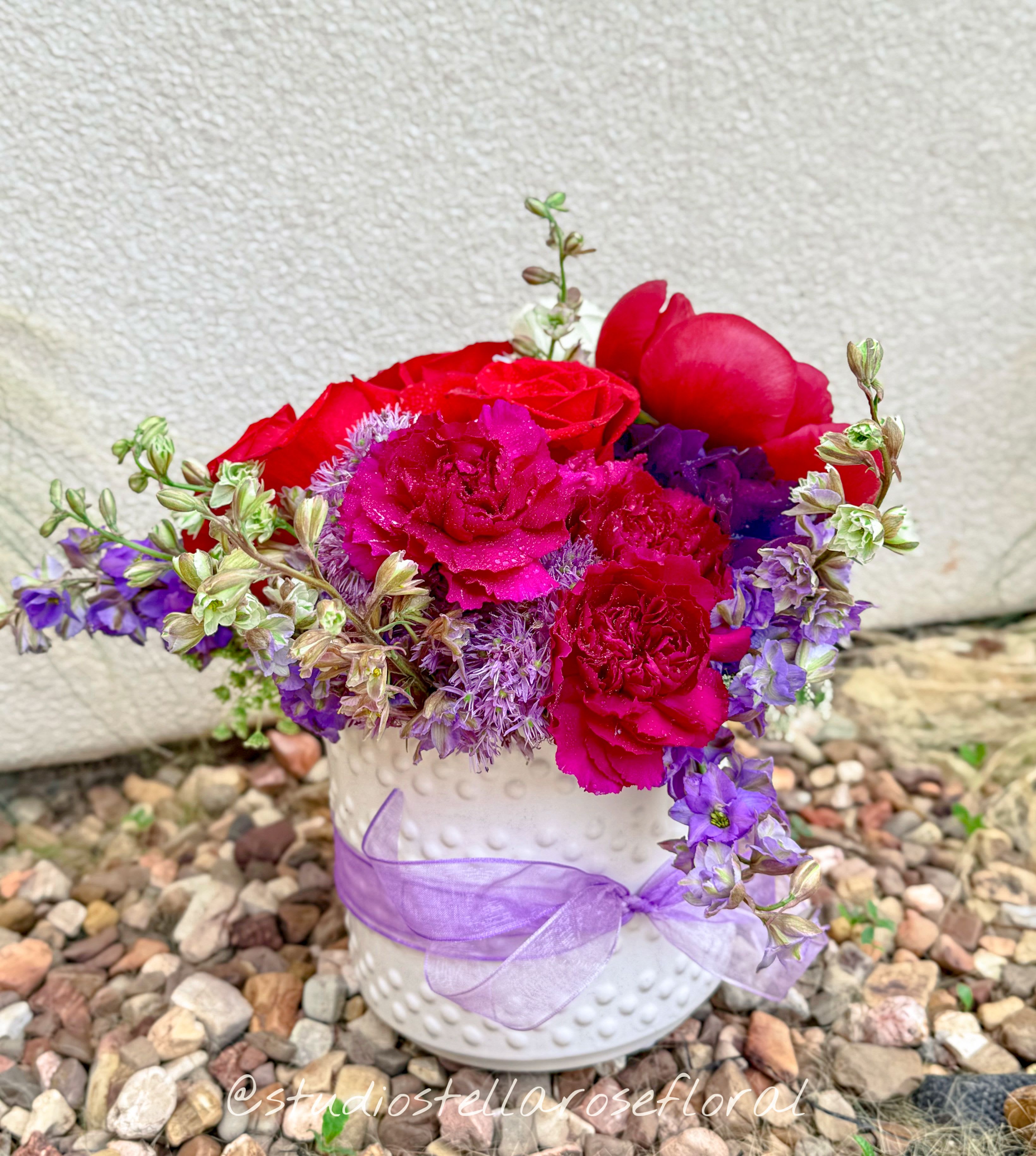 Pink and purple flowers arranged in a white vase with a lavender ribbon.