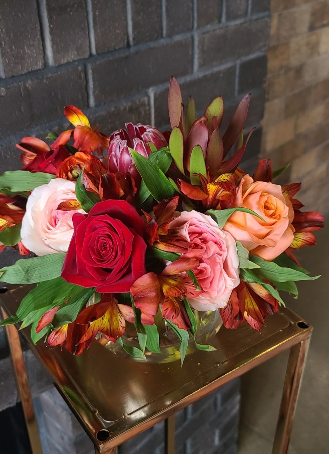 Low arrangement of red, pink, and peach roses with orange alstroemeria in a clear vase on a metal stand