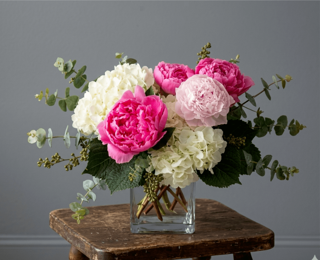 Pretty in Peonies  - a mixture Hot‑pink or light‑pink peonies mixed with creamy hydrangeas, blush accents and eucalyptus in a clear glass vase