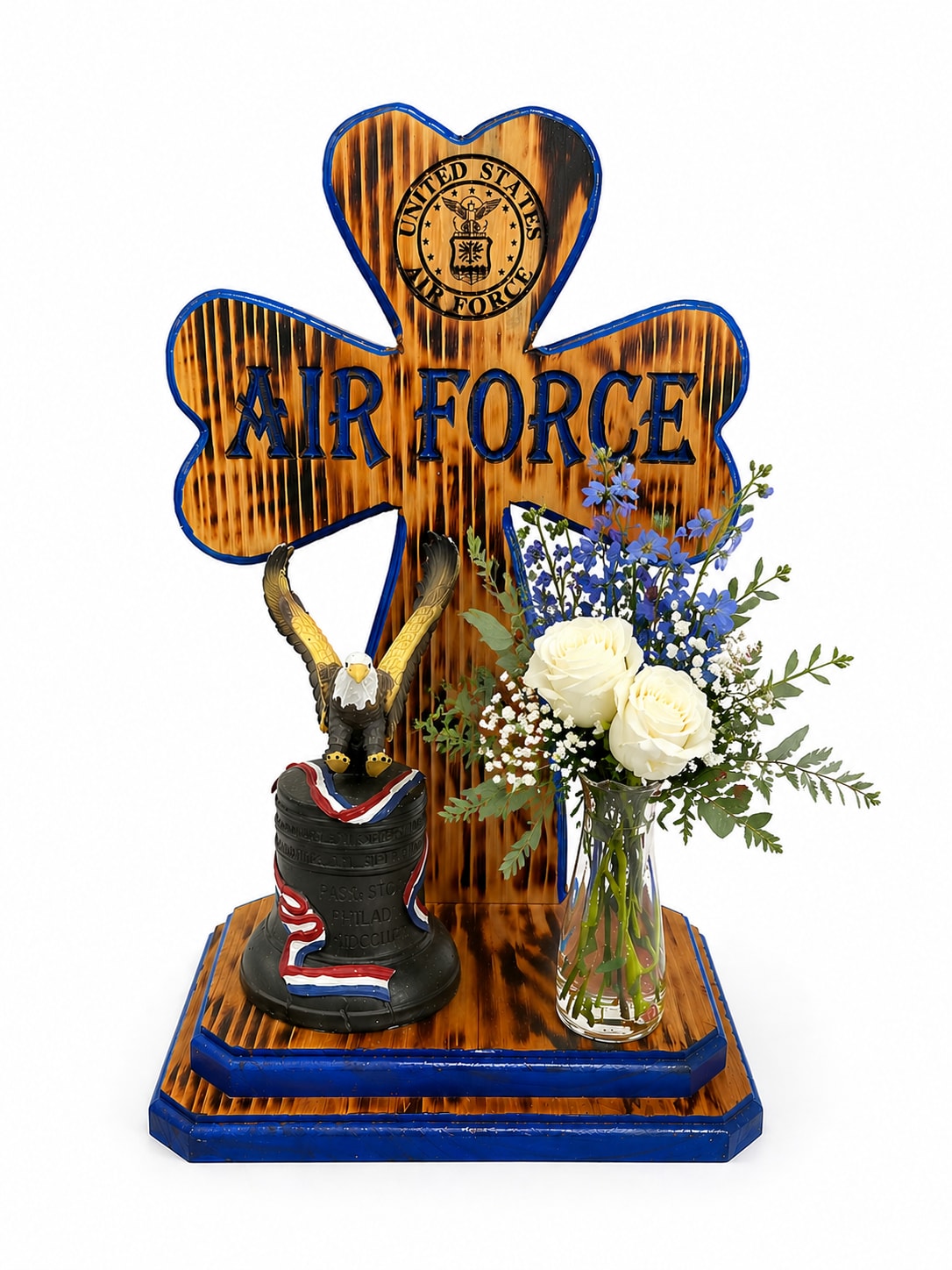 Engraved Wooden Cross - A meaningful and patriotic memorial piece featuring a handcrafted wooden cross engraved with the United States Air Force emblem. The rustic wood finish with bold blue accents creates a strong and honorable tribute.  Accented with a symbolic eagle and bell, this design is paired with a delicate bud vase holding a simple yet elegant arrangement of white roses, soft blue delphinium, baby’s breath, and fresh greenery. The combination of natural elements and patriotic details offers a heartfelt way to honor and remember a loved one who served.