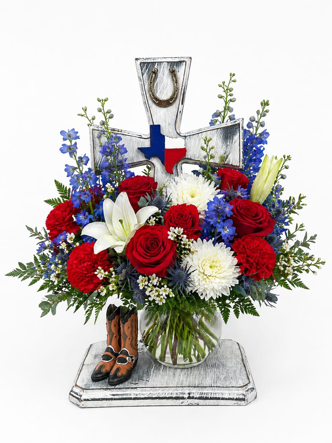 Freedom Cross Tribute - A bold and patriotic floral arrangement designed to honor and celebrate a life with pride and remembrance. This striking piece features a vibrant mix of red roses, white chrysanthemums, lilies, and blue delphinium, beautifully arranged with lush greenery for a full and balanced look.  Set in a clear glass vase, the arrangement is accented by a rustic decorative cross adorned with a Texas emblem and horseshoe, along with a small pair of western boots for a unique, personal touch. The red, white, and blue color palette creates a meaningful tribute, perfect for memorial services or honoring a loved one with strong ties to faith, country, or western heritage.