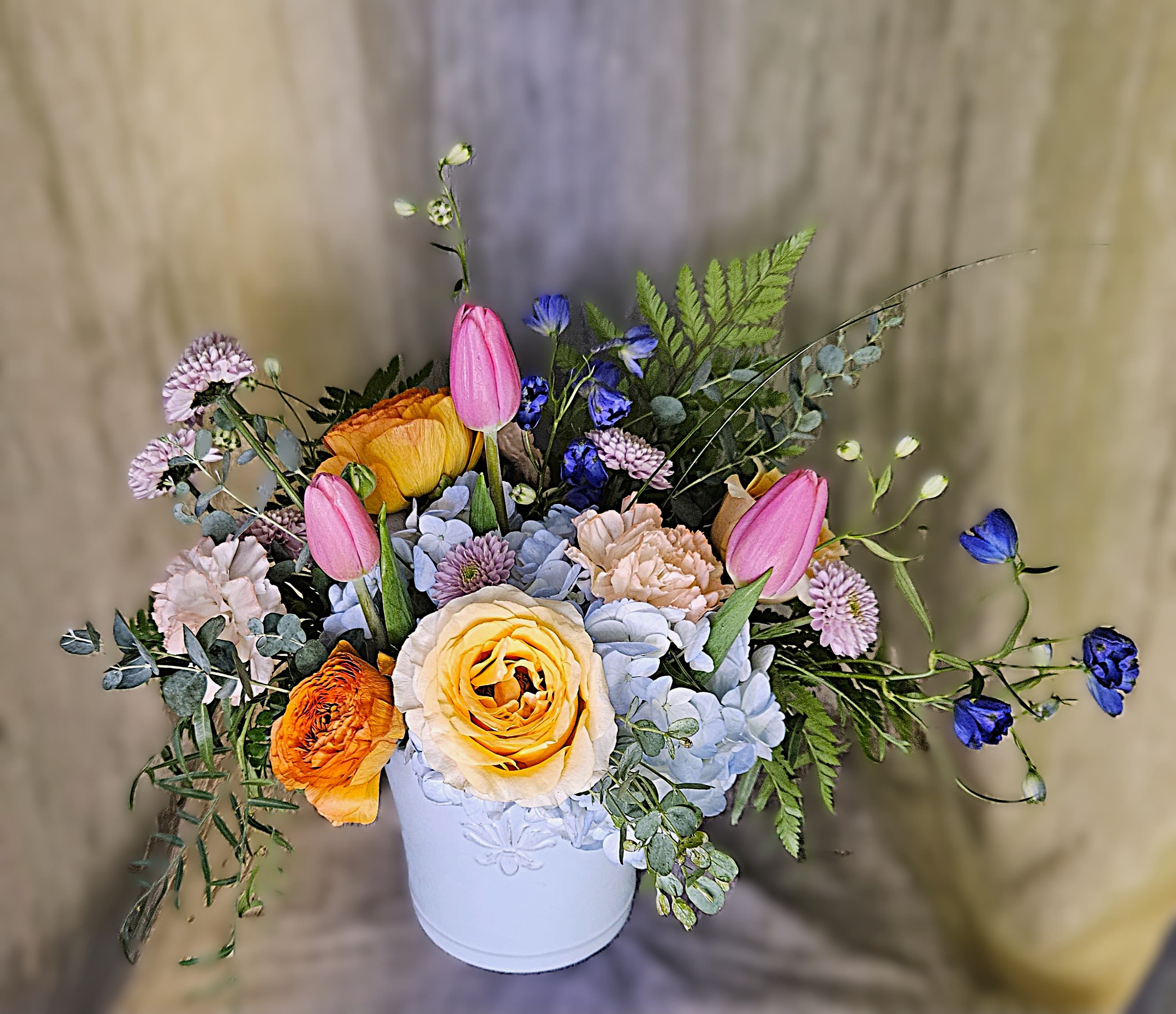 Blushing Blues - PREORDER - A cheerful burst of spring color designed to make Mom smile. Ranunculus, roses, tulips, hydrangea, and soft textured blooms gather loosely in a light‑blue vessel for a relaxed, garden‑style look perfect for celebrating someone special this Mother’s Day.  Available after 5/1/26