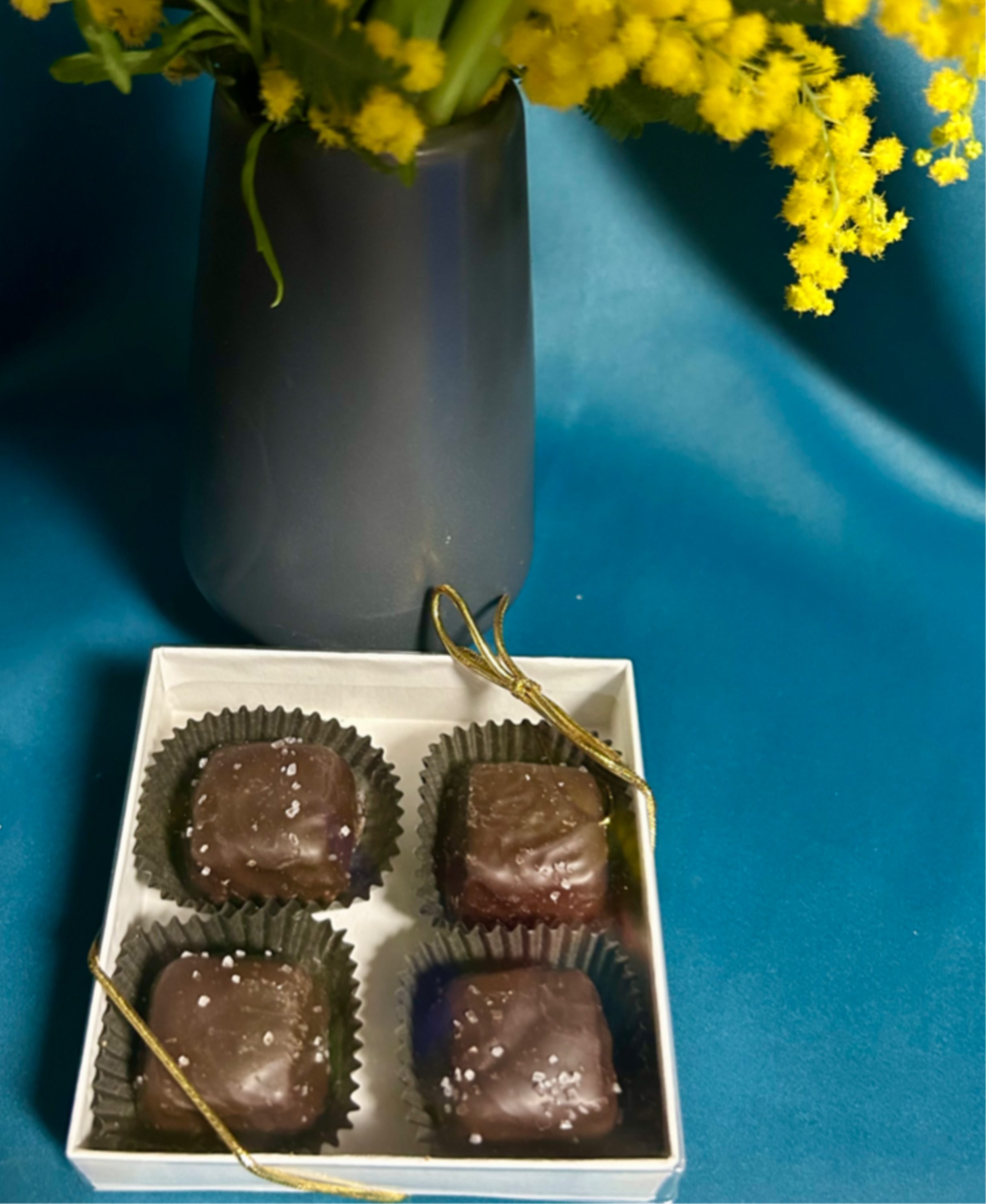 Yellow fluffy flowers in a dark vase beside a white box of four salted chocolate candies