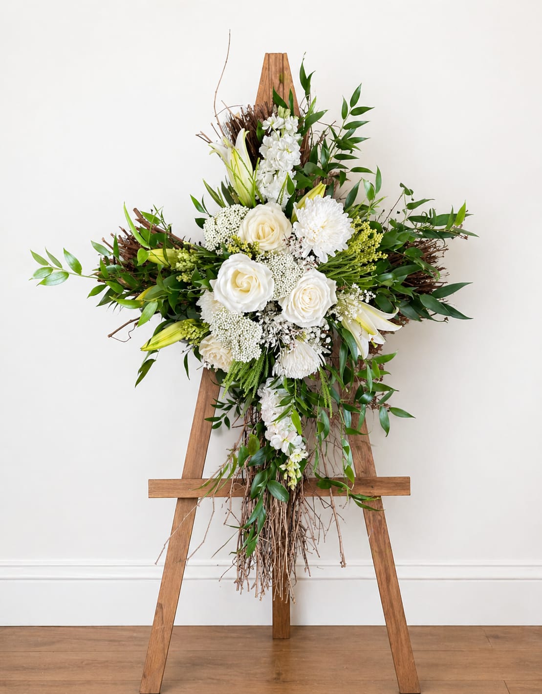 All-White Floral Grapevine Cross - This is an elegant, all-white floral arrangement displayed on a metal green easel. not wood At the center is a lush cluster of white blooms, including:  White roses as the main focal flowers White chrysanthemums and carnations adding fullness and texture White lilies, both open and in bud form, bringing height and grace Delicate filler flowers (like Queen Anne’s lace or similar) for a soft, airy look  The arrangement is surrounded by rich green foliage—long leafy stems and ferns—that create contrast and give it a fresh, natural appearance.  Thin branches are woven throughout the design, extending outward and downward. At the bottom, the branches cascade into a loose, flowing tail, adding movement and a slightly rustic, handcrafted touch.  The overall color palette is white and green, giving the piece a serene, peaceful, and refined look. It feels appropriate for occasions such as memorials, sympathy displays, or formal ceremonies where a calm and elegant presence is desired.