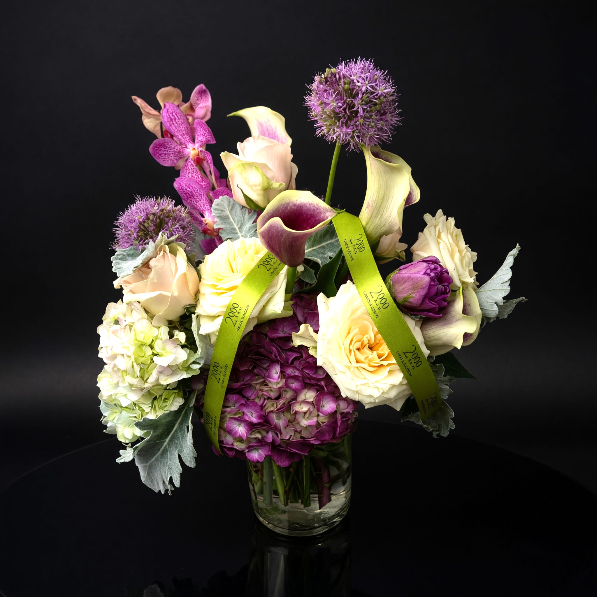 Charlotte - Cream and blush roses, white calla lilies, and lush hydrangea blooms are artfully arranged in a clear glass vase tied with a green signature ribbon, giving both height and fullness from every angle. The palette of purple, white, pink, and touches of yellow plays across textures-silky orchid petals, velvety roses, and crisp calla curves-for a layered, slightly dramatic look that reads as refined yet approachable.