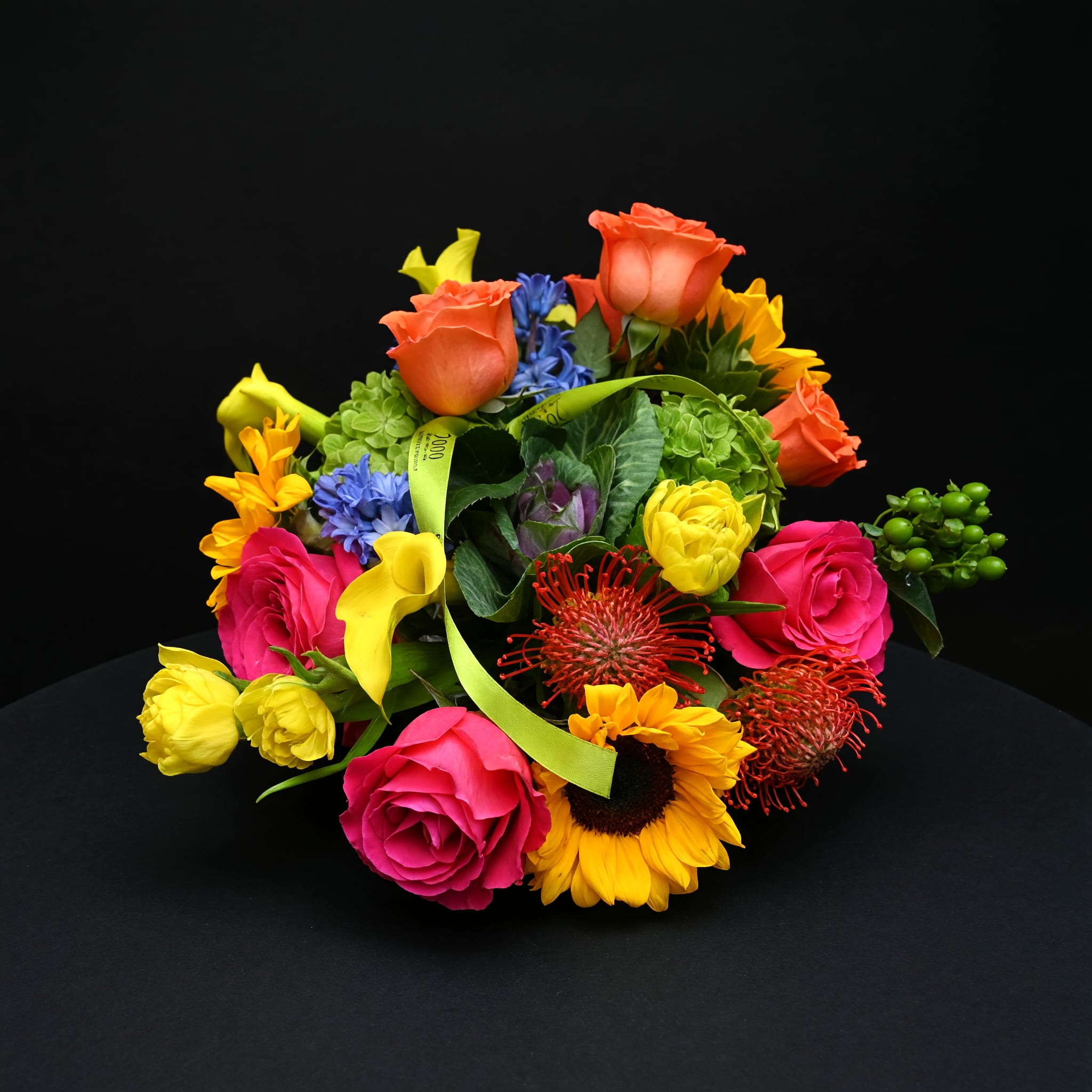 Esme - This arrangement features a mix of roses, mini calla lilies, sunflowers, hydrangea touches and textured filler blooms arranged close to the vase line for a full, layered look. The palette balances warm corals and oranges with hot pink and pops of lime and sky-blue, while a lime-green ribbon woven through the center adds a tailored, gift-ready finish. Compact and low in stature, it sits beautifully on a table or desk without blocking conversation.