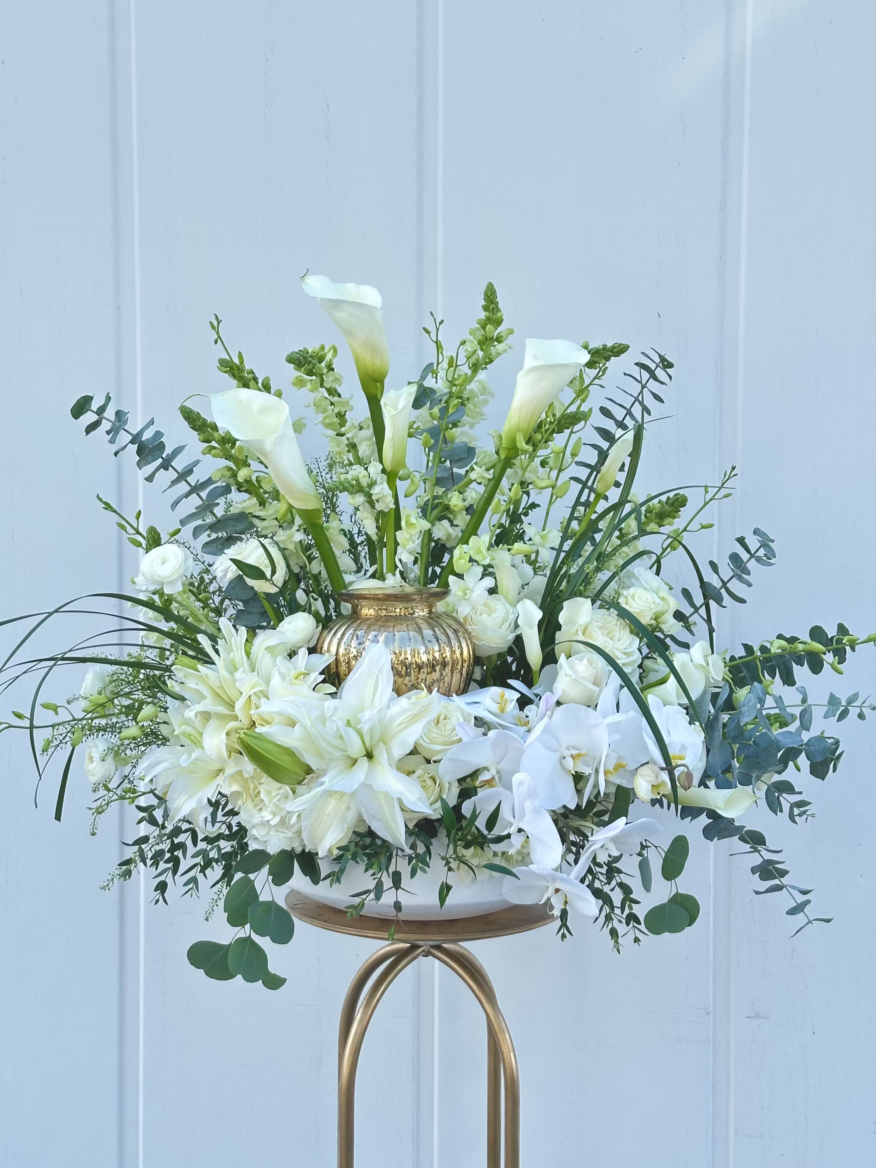 CREMATION TRIBUTE  - An airy, elegant sympathy flower arrangement built to honor a life with quiet luxury - this cremation tribute features white calla lilies, phalaenopsis orchids, roses and lilies arranged with soft eucalyptus-like foliage in a reflective gold vase.  