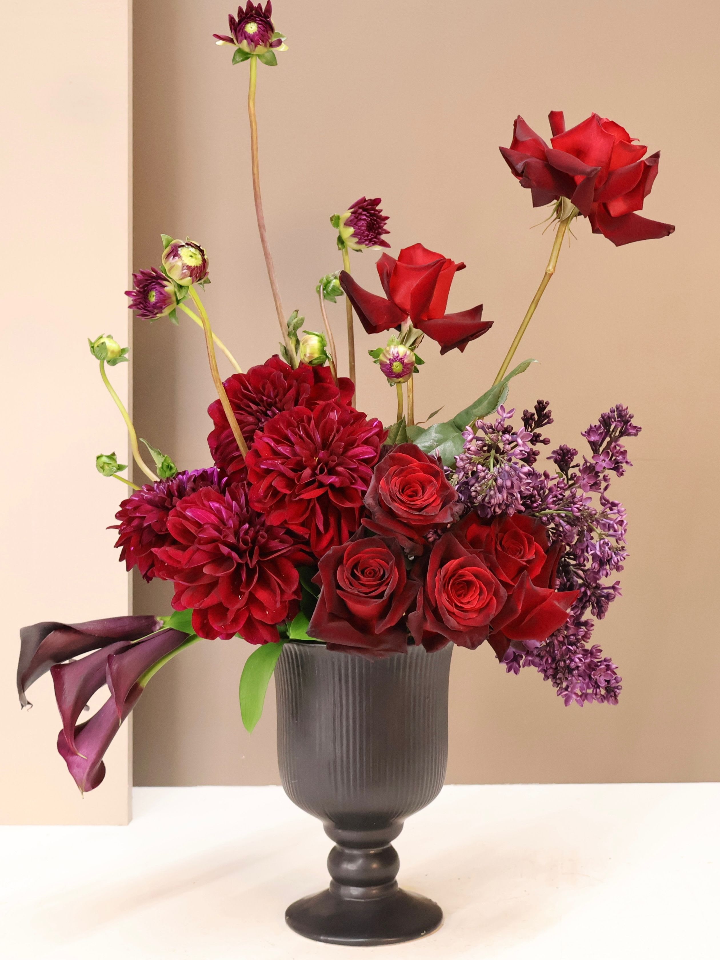 Burgundy Luxe - A striking arrangement of deep Burgundy Roses and lush Dahlias accented with sculptural Calla Lilies and fragrant Lilac. Designed in a modern pedestal vase, this bold composition brings drama and a bit of whimsy.