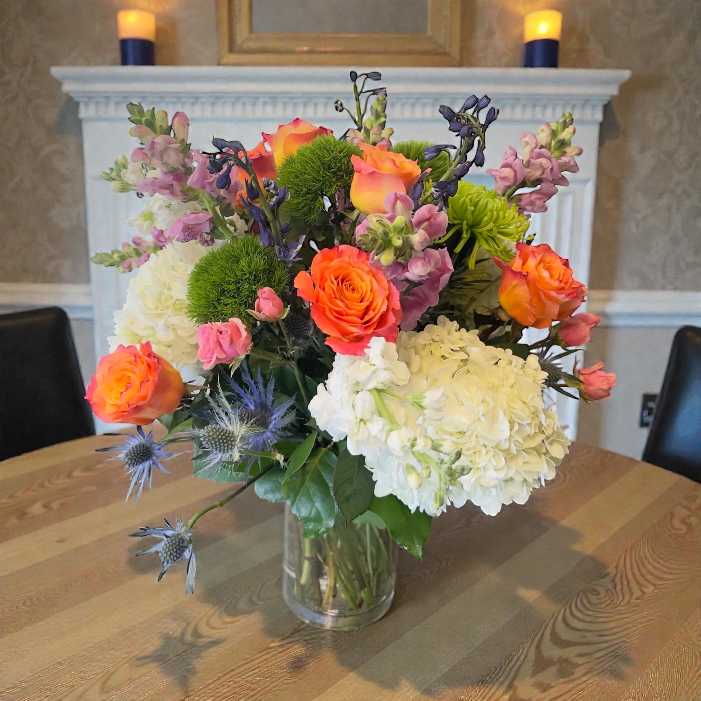 Solara Bloom Symphony #7 - A bold cluster of orange roses and cloudlike white hydrangeas makes an immediate, joyful impression-perfect as a Mother's Day or birthday flower arrangement that will make the person you're thinking of feel celebrated. This rounded garden-style bouquet reads as bright and abundant, an ideal choice for anniversaries, housewarmings, graduations, or any "just because" moment when you want someone to smile.  This mixed bouquet combines orange roses, white hydrangeas, purple snapdragon-like spikes, delicate spray roses and daisies, plus small blue thistle-like accents for contrast, all arranged densely in a clear glass vase. The varied textures-soft hydrangea fluff, sculpted rose petals, upright snapdragon spikes, and spiky blue accents-create depth and a lively color story of pink, white, orange, blue and purple that suits both spring and summer gifting. The vase gives it a tidy, ready-to-display presence while keeping the overall look relaxed and garden-fresh.  Thoughtful for many occasions-Father's Day, Thanksgiving, Get Well, Thank You, Graduation, Prom, Retirement, or simply Thinking of You-this arrangement reads as warm and sincere rather than overly formal. For best enjoyment, keep the water fresh, trim stems slightly every few days, and place away from direct heat to extend bloom life; it arrives ready to brighten a table, entryway, or bedside with minimal fuss.