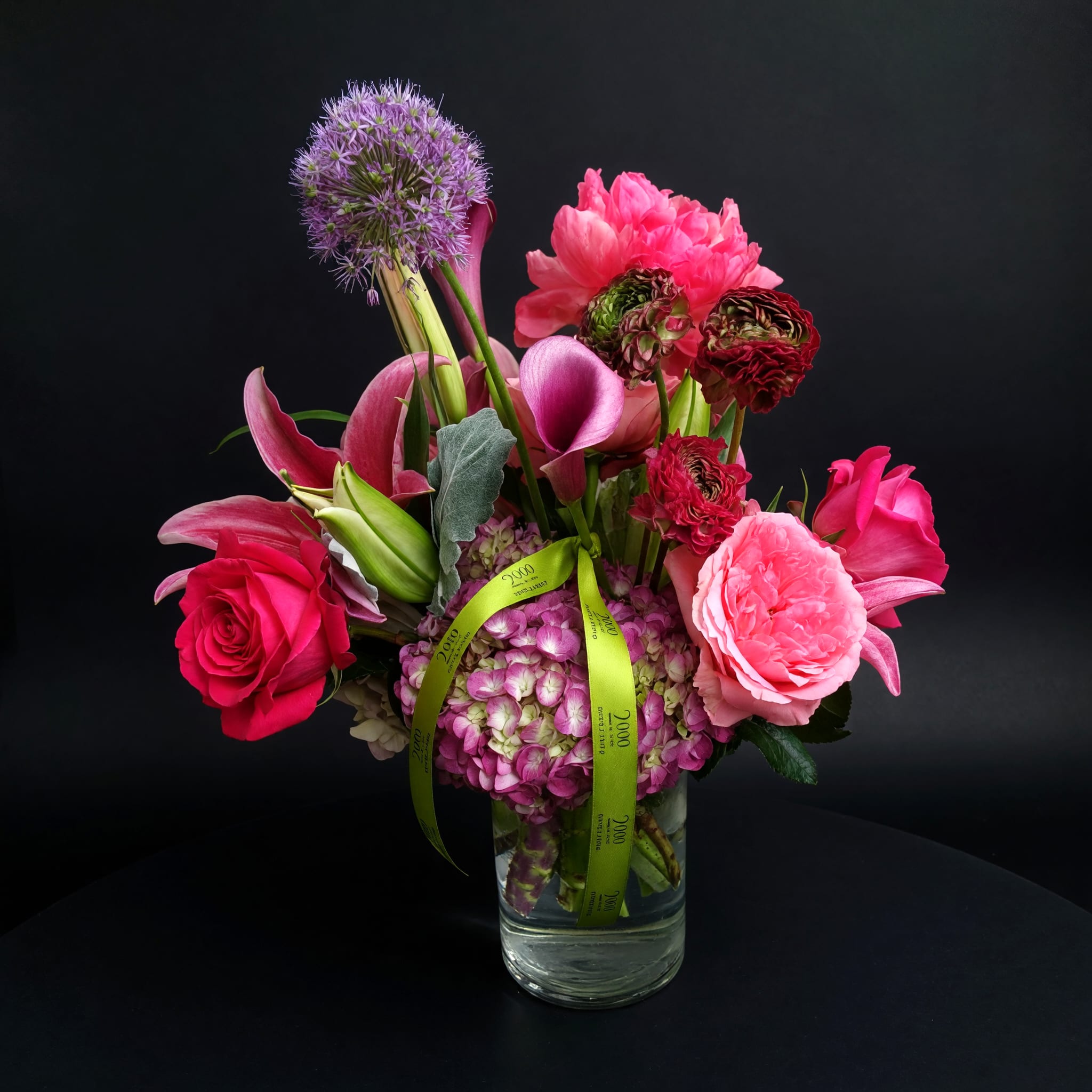 Juliet - Large hydrangea heads anchor the composition while fragrant lilies and graceful calla lilies add height and elegant sweep. Classic roses and ruffled ranunculus fill the mid layers for a soft, rosy texture, and a spherical allium provides a sculptural purple pop that catches the eye. The palette of pink, purple, red and white is intentionally feminine and vibrant, with varying shapes and heights that create a full, garden-like feel.