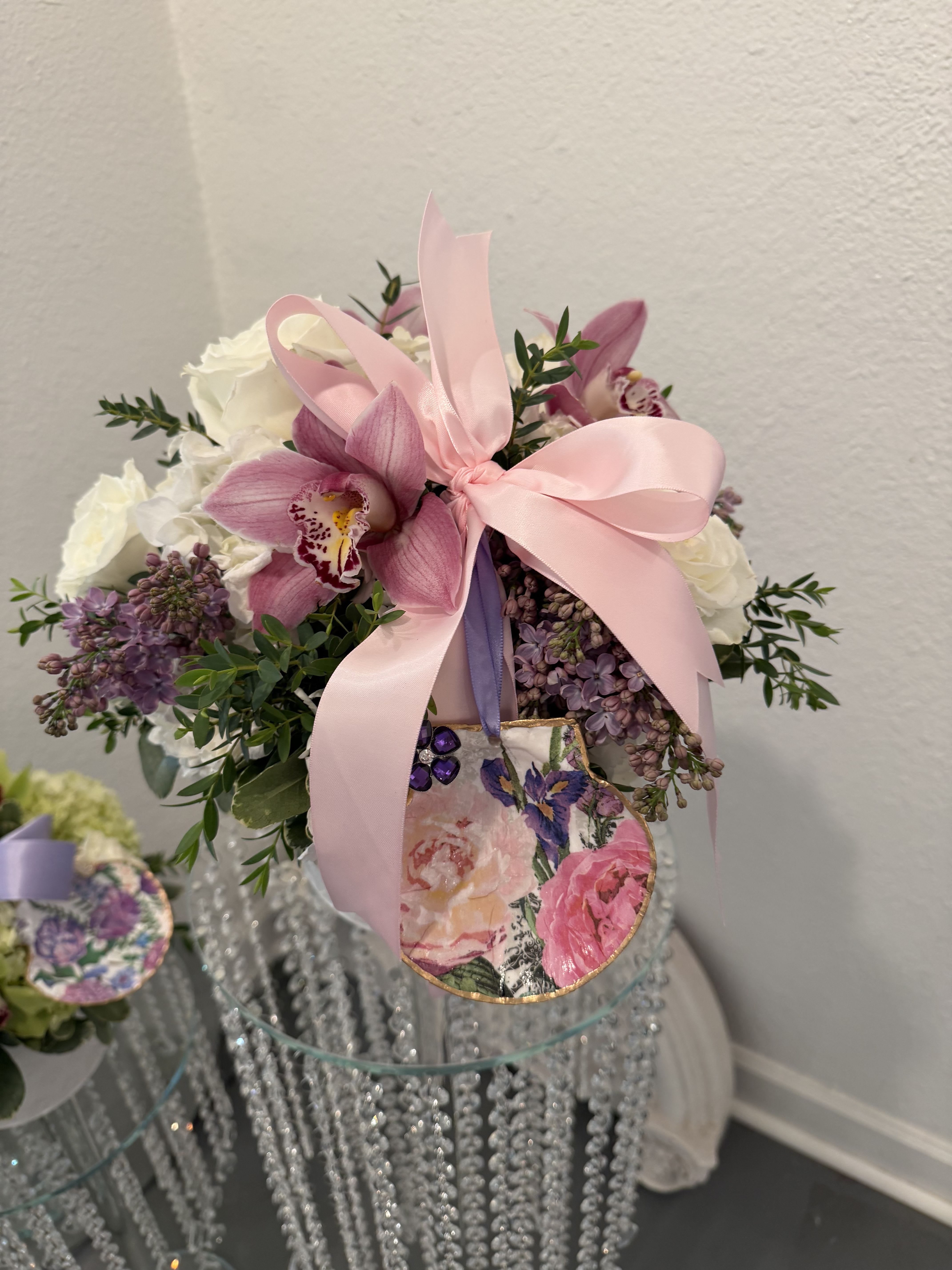 Cherry On Top Bouquet  - A luminous mix of pink, white, and purple florals makes this flower arrangement feel like a sweet celebration-perfect as a birthday bouquet, Mother's Day gift, or anniversary surprise. The rounded, compact design places a pale pink satin bow  and decoupaged shell front and center so the person you're thinking of will see the sentiment instantly; soft lavender tones and fresh greenery lend a romantic, feminine presence that reads as both joyful and elegant.  Built to impress on a tabletop or event pedestal, the Cherry On Top Bouquet pairs orchids, roses, lilac and hydrangea in a white ceramic container. Finishing touches include decorative floral decoupaged shell, pink and lavender ribbons, details that make the arrangement feel gift-ready and polished.  Whether sent to honor a milestone, brighten a mom's day, or elevate a special celebration, this arrangement delivers a feeling of being truly seen and cherished. Order with confidence for same-week delivery where available; stems arrive ready-to-display in their vase with simple care-fresh water and a clean vase will keep blooms looking their best. 