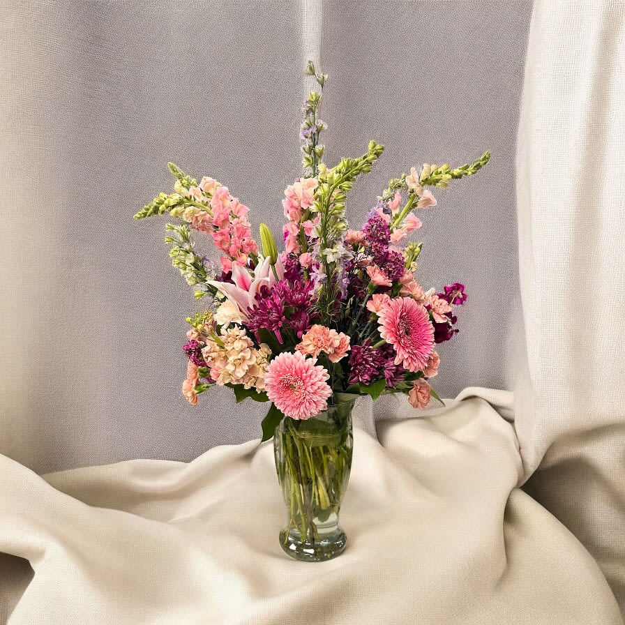 Veloura Garden Whisper #17 - Lavender/pink snapdragons rising above soft pink gerbera daisies give this garden-style flower arrangement a breezy, whimsical silhouette-perfect as a birthday bouquet or a cheerful spring gift. The Veloura Garden Whisper #17 is an everyday floral arrangement that feels thoughtful without fuss, a bright way to say thank you or simply let someone know you're thinking of them.  A loose, airy mix of pink, purple, white, and green includes gerbera daisies, snapdragons, carnations, lilies, and chrysanthemums arranged in a clear glass vase. Tall spires create layered height while fuller blooms add texture and volume; the palette balances pastel romance with lively color notes. The simple glass vase keeps the focus on the flowers' shapes and soft hues for a fresh, modern presentation.  Send this to a friend, family member, or coworker who deserves a lift-the kind of bouquet that makes her feel noticed and celebrated. Vase-ready and low on formality, it's easy to care for: keep in fresh water, recut stems if needed, and display away from direct heat for the longest enjoyment.