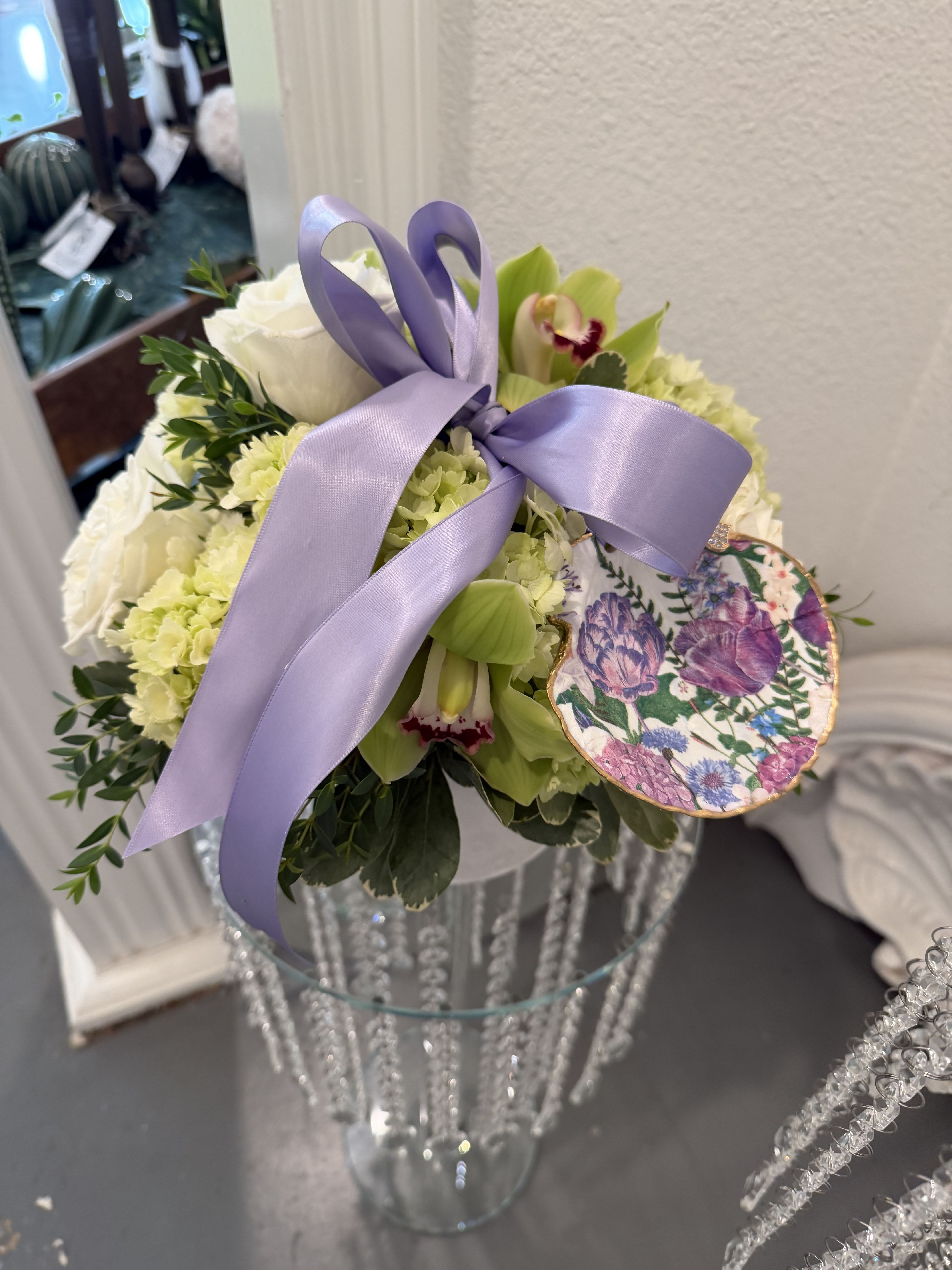 In The Lime Light Bouquet  - Soft whites and pale green florals take center stage in this In The Lime Light bouquet, a flower arrangement made to make someone feel quietly celebrated. Designed as a birthday bouquet, thank-you gift, or a springtime surprise, the calming mix of white, green, lavender and touches of pink reads both romantic and elegantly modern - the kind of arrangement that says "you matter" without saying a word.  A decorative floral decoupaged shell and neat ribbon treatments add a polished, gift-ready look.  Perfect for Mothers Day, an anniversary, a bridal shower or to brighten a bedside or entry table, this arrangement suits both everyday gifting and more formal celebrations. Sizes and hard goods vary by presentation-choose the single glass-vase style for a compact, tabletop statement or the pedestal pair for added height and event flair-each ready to delight the person you're thinking of with lasting beauty and refined detail.