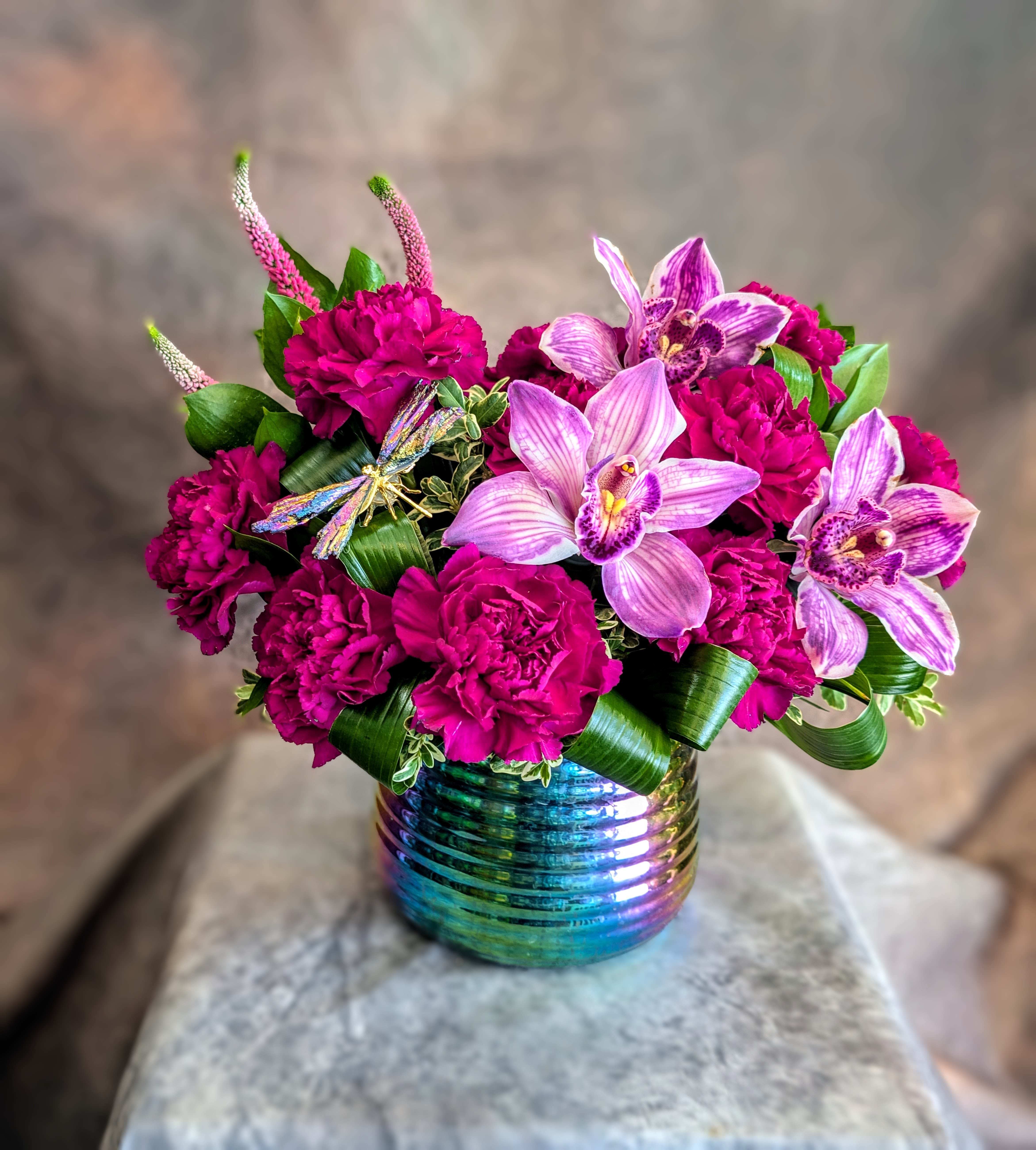 Pixie - Pixie is a vibrant, whimsical arrangement that feels like a little spark of magic captured in bloom. Lush plum carnations create a rich, velvety base, their deep jewel tones adding depth and drama, while variegated purple orchids rise above with elegant movement and intricate striping, adding a striking, artful contrast. Set in a radiant, iridescent ceramic container that shimmers with shifting hues, Pixie catches the light from every angle, making it a true statement piece. Curled tropical greens add structure and a modern, sculptural feel, while a delicate dragonfly accent brings a touch of enchantment—like a fleeting moment in a secret garden. Handcrafted in Las Vegas by Windmill Floral Expressions, each Pixie arrangement is made fresh to order using premium blooms and our signature artistic design style. No two are exactly alike, ensuring a one-of-a-kind creation every time. Perfect for birthdays, celebrations, or anyone who loves bold color and a little magic, Pixie is available for same-day flower delivery in Las Vegas and Henderson.cc