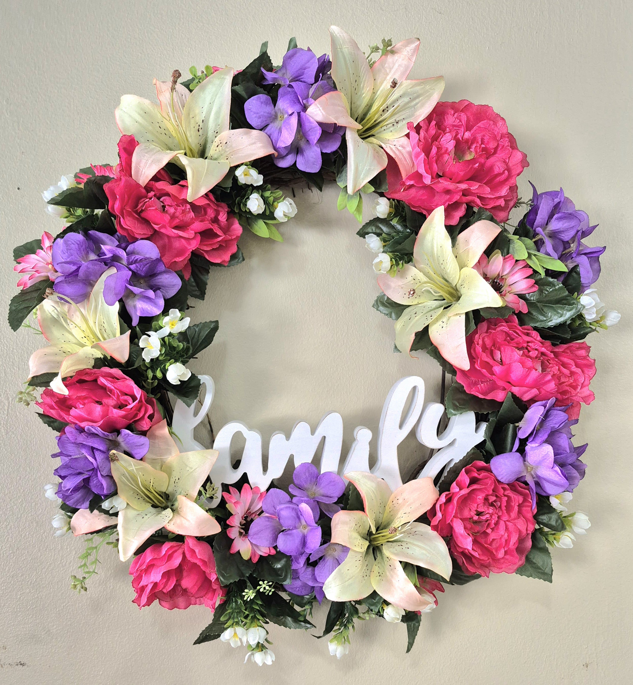  Family "Silk" Grapevine Wreath  - A full-circle silk wreath layered with bright pink peony-style blooms, pale peach and cream lilies, and soft clusters of purple florals - this family "Silk" wreath greets visitors with a gentle, homey charm perfect for a birthday, housewarming, or just because. The white wooden "family" script in the open center turns the piece into a warm statement for a front door, entryway, or mantle, promising a cheerful first impression whether it's spring, summer, or any day you want to celebrate home.  Crafted from long-lasting silk blossoms, the 20" wreath combines textured petals and tiny white filler flowers for depth and visual movement around the ring. The mix of pink, purple, white, and peach hues keeps the look fresh and seasonal without feeling limited to one occasion. Lightweight and ready to hang on a door or wall, it's an easy way to brighten a new home, mark a birthday, or bring springtime color indoors - a thoughtful gift that will continue to welcome family and friends year-round.