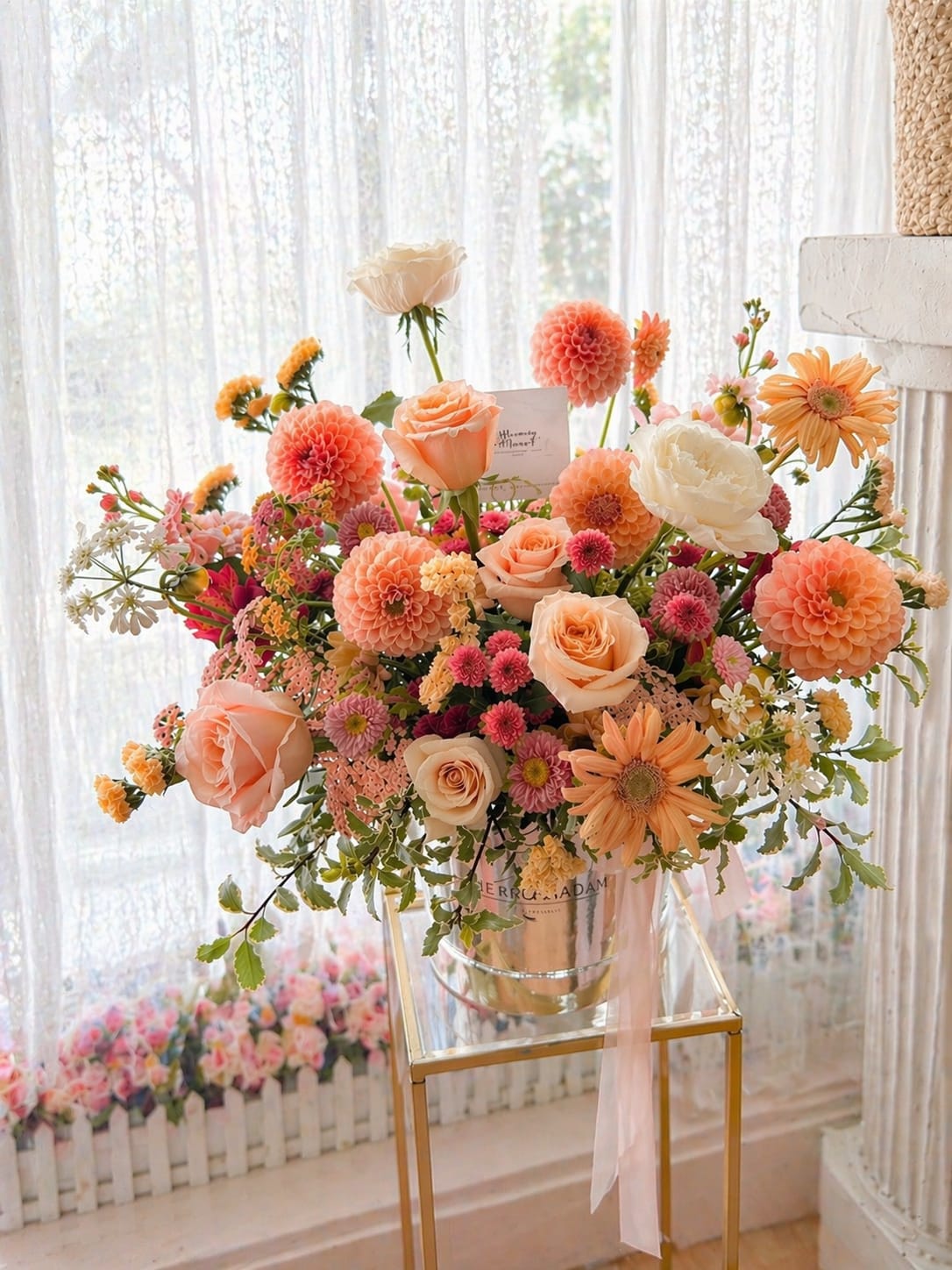 Peach Blush Garden Arrangement - A soft, garden-inspired arrangement in warm peach and blush tones, designed to feel natural, airy, and effortlessly elegant. Featuring premium roses, dahlias, spray blooms, and delicate seasonal accents, this piece brings a fresh, romantic charm to any space.  Styled in a modern glass vase with flowing ribbon detail, it’s perfect for birthdays, celebrations, or simply brightening someone’s day with a touch of gentle beauty.  Each arrangement is handcrafted with the freshest blooms available. Flower varieties may vary slightly while maintaining the overall color palette and style.