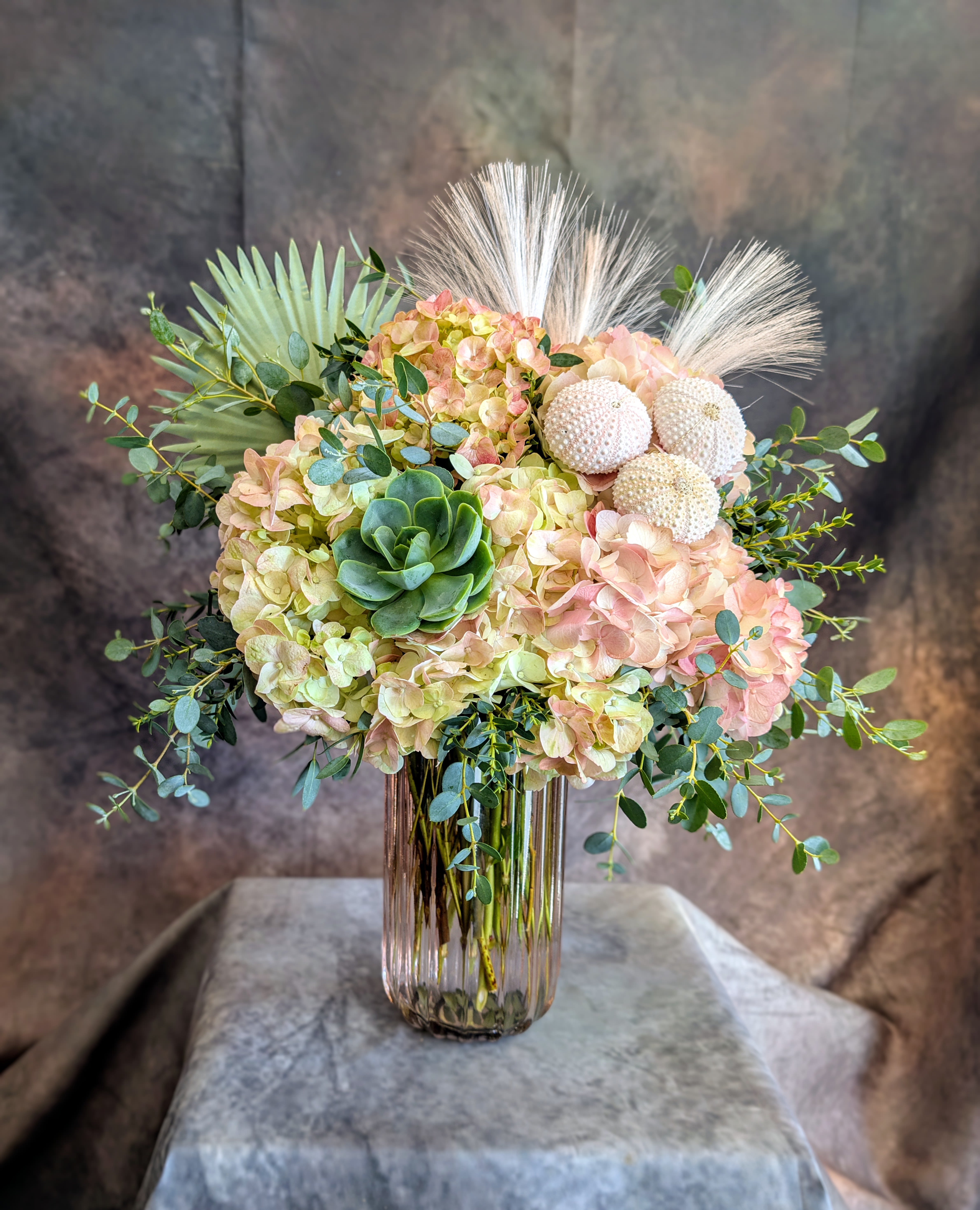 Mia - Mia is a softly layered arrangement that blends natural texture with understated elegance. Cloud-like hydrangeas in delicate tones of blush, antique green, and warm peach create a lush, romantic base, while a sculptural echeveria succulent adds a modern botanical touch.  Accents of airy pampas grass and natural sea urchin shells bring in a coastal, textural element, giving Mia a serene, sun-washed feel—like a quiet moment where desert meets sea. Wisps of fresh greenery add movement and softness, completing the design with an organic, effortless flow.  Designed in a clear glass vase that showcases the natural beauty of each stem, Mia is both refined and relaxed—perfect for everyday luxury, thoughtful gifting, or adding a calming, elevated presence to any space.  Handcrafted in Las Vegas by Windmill Floral Expressions, each arrangement is made fresh to order with premium blooms and our signature artistic style. Available for same-day delivery in Las Vegas and Henderson.