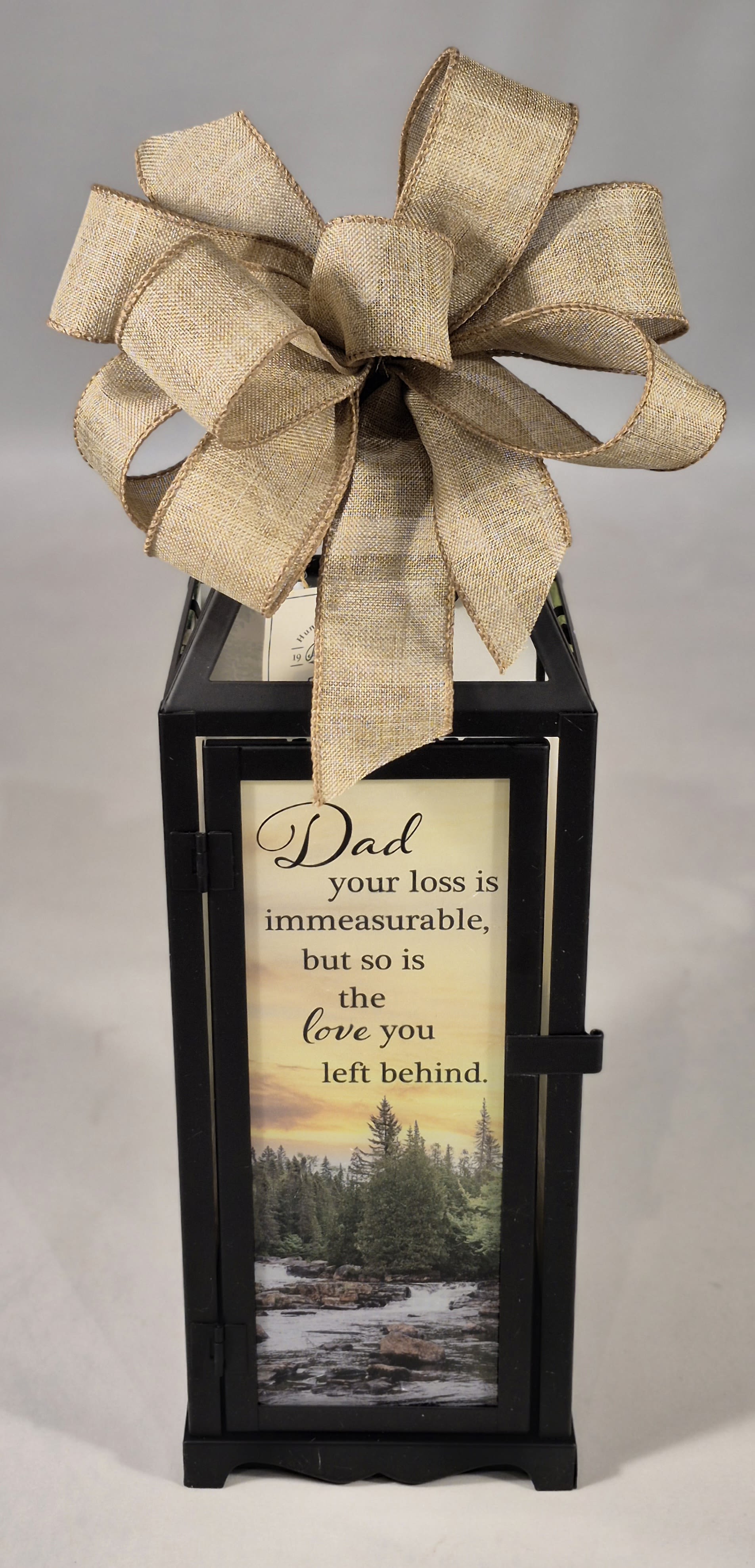 Dad Panoramic Lighted Sympathy Lantern  - The heartfelt sentimental message and scenic nature imagery on this lantern for the loss of a loved one. The message reads: "Dad, your loss is immeasurable, but so is the love you left behind", making a lovely remembrance piece.  This beautiful lantern measures 15.5"H x 4.75"W.   This lantern features a flameless LED candle with a beautiful flickering appearance for a warm ambience. When switched on, the light runs on an automatic timer of 6 hours on, 18 hours off. Lantern requires 3 AAA batteries included. Choose deluxe if you would like to add a Silk arrangement on lid of lantern.