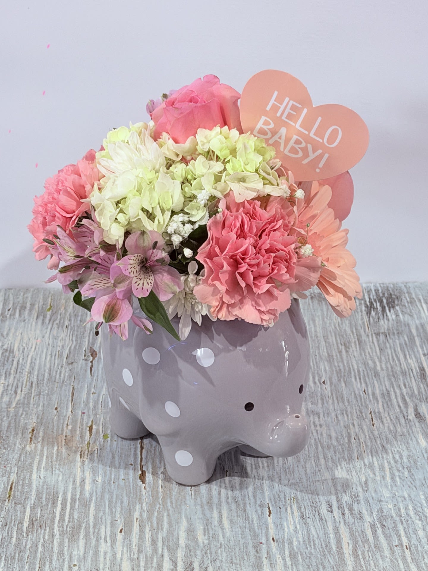 Welcome Baby Girl Elephant - Soft pink carnations and pale hydrangeas mingle with white daisies and lavender alstroemeria in a compact, rounded bouquet made to welcome a new baby girl or brighten a recovery room. This welcome baby flower arrangement reads as an instant nursery accent: the glossy lavender ceramic elephant planter with white polka dots and a peach heart sign that says "HELLO BABY!" create a playful, gift-ready presentation perfect for New Baby visits or a cheerful Get Well surprise.  The palette of pink, white and lilac feels gentle and uplifting; the textures range from the ruffled carnations and full hydrangea heads to the delicate alstroemeria and crisp daisy centers. The arrangement is designed to sit on a dresser or bedside table - a decorative, compact piece rather than a tall vase display - and arrives ready to enjoy in its keepsake elephant planter. It's an easy, thoughtful way to tell the new mom or someone recovering that you're thinking of them.