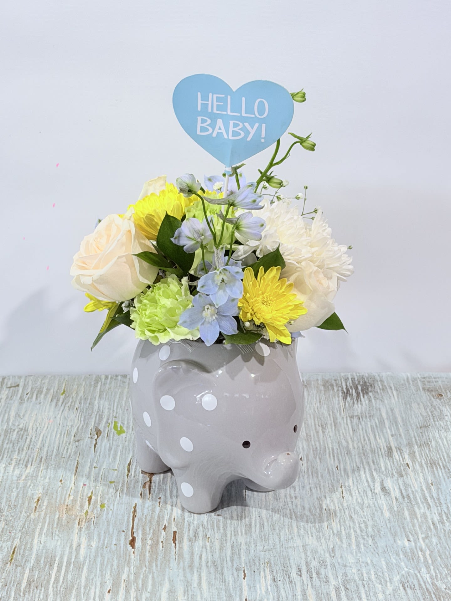 Welcome Baby Boy Elephant - Soft pastels and a playful gray elephant make a joyful hello for a newborn - perfect as a new baby or baby shower gift. This welcome baby boy flower arrangement feels nursery-ready: the compact, rounded design and blue heart sign that reads "HELLO BABY!" announce the occasion while keeping the mood light enough for a get well bedside pick-me-up.  Creamy white roses mingle with yellow and white chrysanthemums, pale blue delphinium, and a soft green bloom for gentle texture and cheery contrast. Everything is planted in a gray ceramic elephant container with white polka dots and finished with a heart-shaped baby sign; the size and low, rounded silhouette suit a hospital tray, nursery shelf, or small entry table.  Give this to new parents to celebrate their little one or send it to brighten recovery time - it's the kind of arrangement that makes someone feel seen and celebrated. Care: keep blooms in a cool spot away from direct sun and replenish vase water as needed to extend freshness.