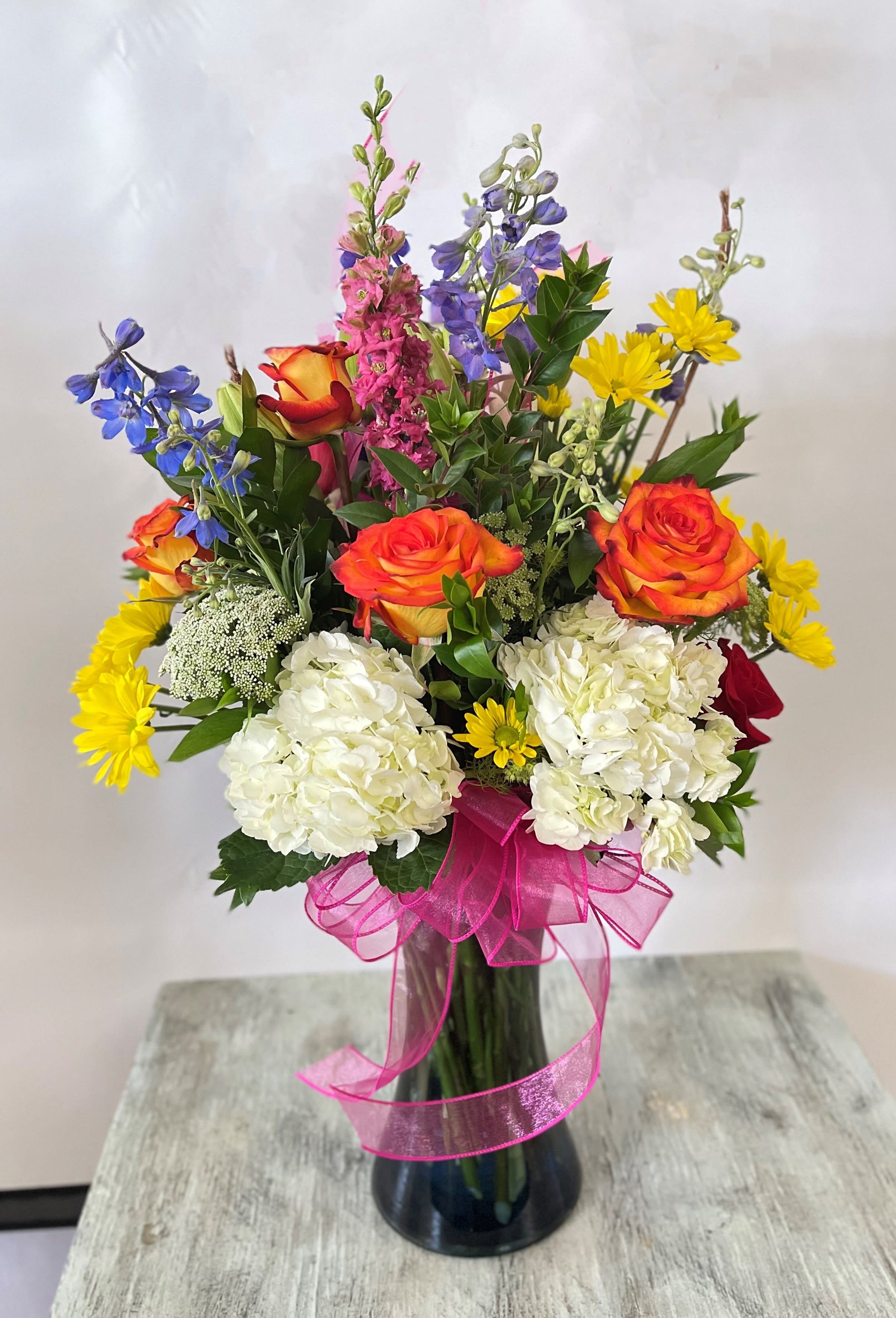 Mixed bouquet of roses, hydrangeas, and daisies in a glass vase with a pink ribbon