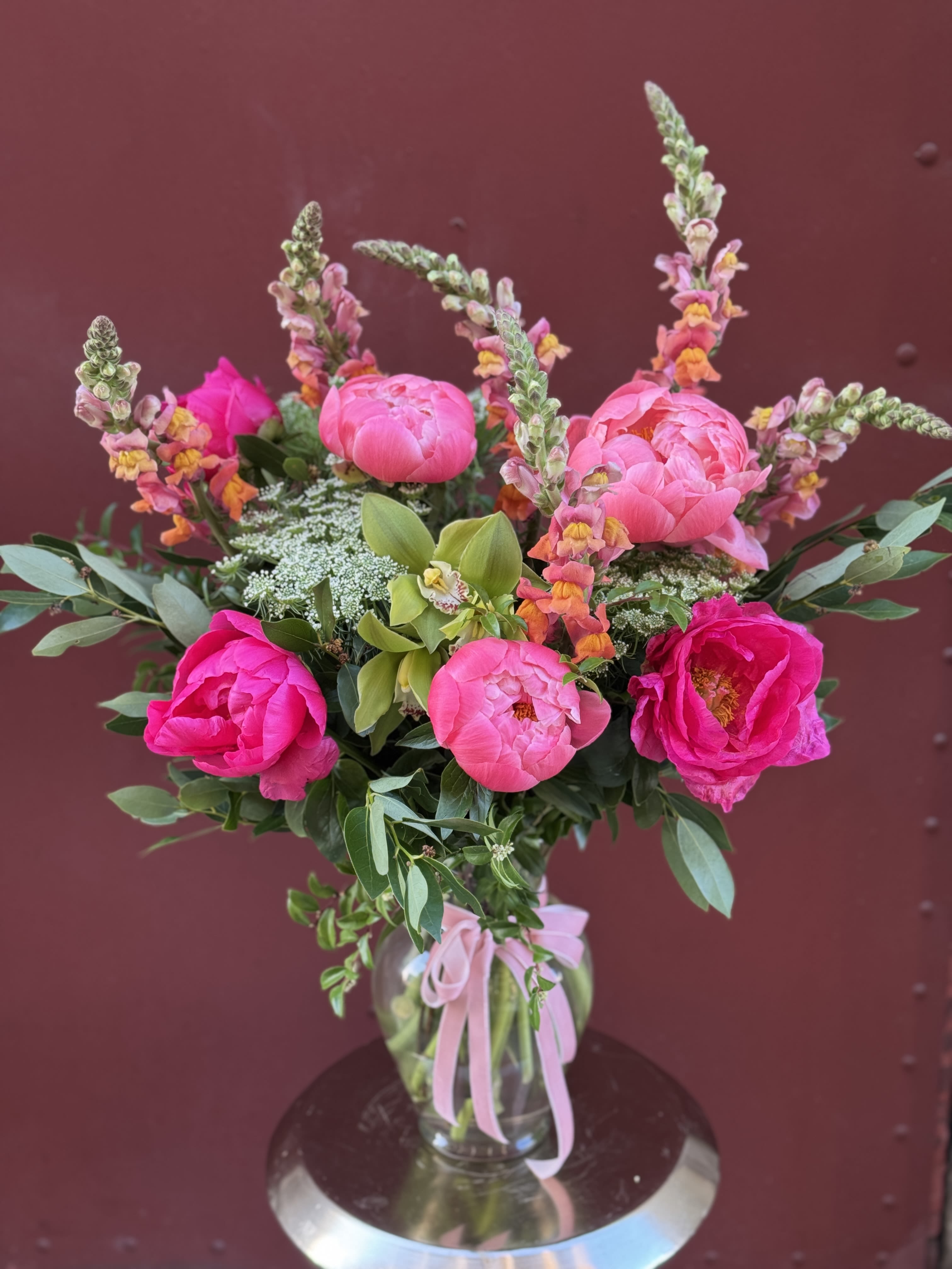 Pink peonies and snapdragons arranged in a glass vase with a pink ribbon