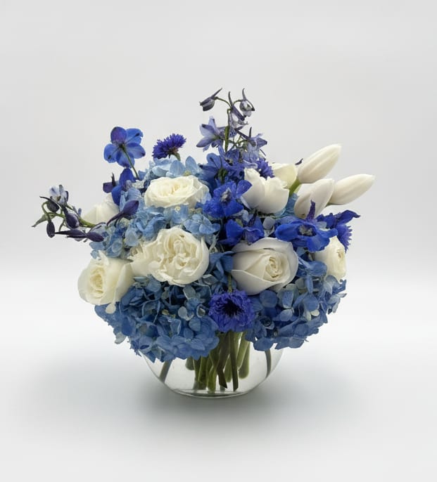 BLUE LAGOON - A cool, rounded cluster of blue hydrangeas and creamy white roses makes this flower arrangement feel like a gentle ocean breeze - perfect as a birthday bouquet, anniversary surprise, or a serene Mother's Day gift. The blue-and-white palette with soft purple-blue accents reads fresh and refined, so the person you're thinking of will feel calm, cared for, and truly seen.  Dense blue hydrangeas anchor the design, paired with white roses for clean lines and soft contrast; small purple-blue accent blooms and delphinium add texture and depth throughout. The arrangement is presented in a clear glass vase for a modern, unfussy finish and a compact, rounded silhouette that suits tabletops or bedside dressers.  Ideal for spring and summer celebrations, new baby welcomes, or Admin Professionals' Week, this is the kind of bouquet that feels appropriate for both milestone moments and thoughtful everyday gestures. Standard sizing fits most spaces; to keep blooms looking their best, refresh vase water every 2-3 days and trim stems slightly when you change the water.Add a little something extra, gourmet chocolates, hand poured candles. See all our gift selections under the thoughtful gifts tab. Or call the shop and let us know what you are looking for and we will do the shopping for you.
