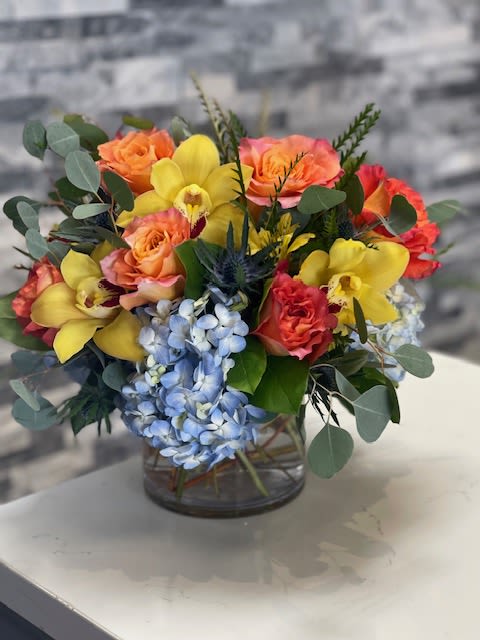 Short arrangement of orange roses, yellow orchids, and blue hydrangeas in a clear glass vase.