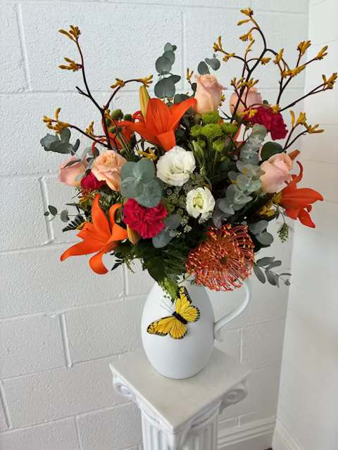 Porcaline Blooms - Bright orange lilies and blush roses take center stage in this lively flower arrangement, making it an eye-catching choice for a birthday bouquet, a cheerful thank-you gift, or an everyday pick-me-up. The mix of orange, pink, white and green tones reads warm and autumn-leaning, so the person you're thinking of will feel genuinely celebrated the moment these blooms arrive.  Hand-gathered with white roses, red carnations and a round orange pincushion-style bloom, the design uses silvery eucalyptus and branching yellow accents for a loose, airy structure that adds height and movement. Everything is arranged in a matte white pitcher vase with a playful yellow butterfly decoration-perfect for displaying on a kitchen table or mantel as a keepsake after the blooms fade.  This arrangement suits someone who loves bold color and a slightly rustic, expressive style; it's versatile for birthdays, thank-yous, or brightening a room. Standard care-fresh water and a cool spot away from direct sun-will help the lilies and mixed bouquet elements last as long as possible.
