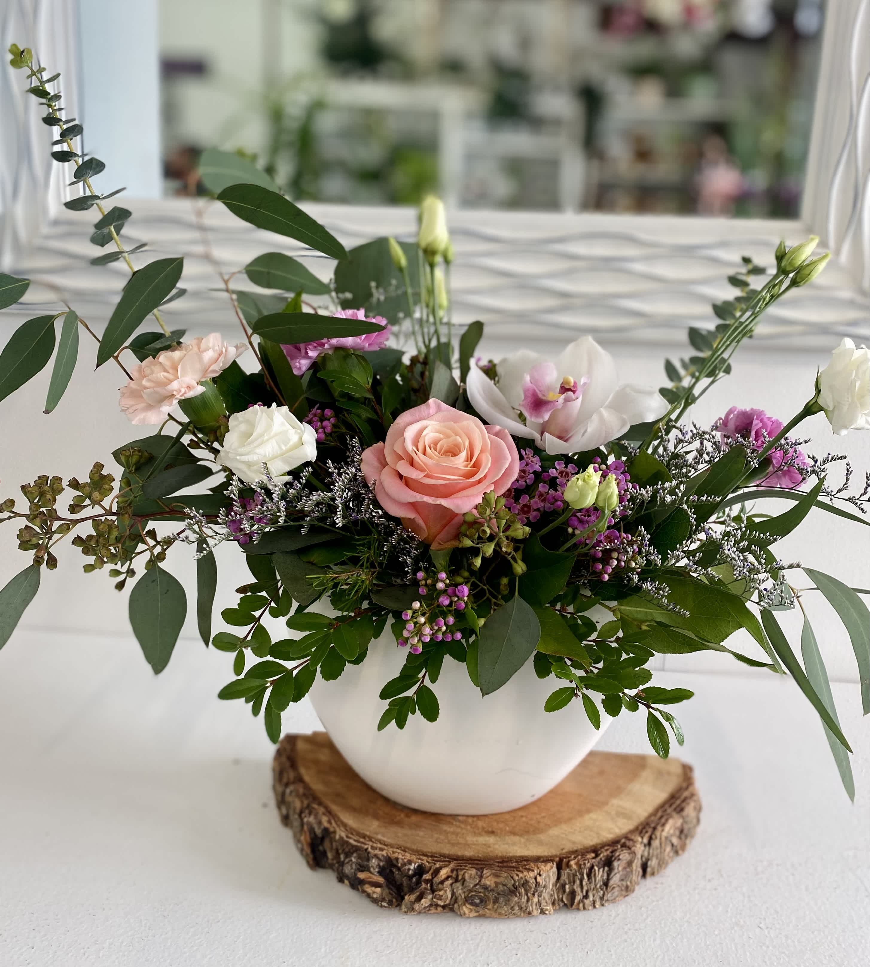 Low pink and white flower arrangement with roses, orchids, and lisianthus in a white bowl on a wood slice base