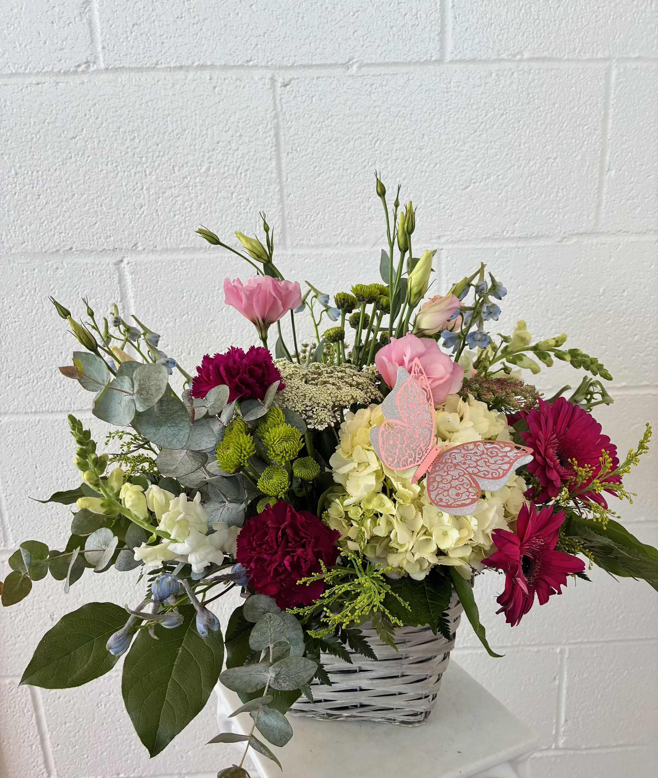 Blooming Devotion - A soft spring palette of pink, white, purple, and green blooms creates an uplifting first impression-perfect for a birthday bouquet, thank-you arrangement, or just because. This mixed flower arrangement in a white woven basket feels both feminine and fresh; the person you're sending it to will notice the bright pink gerbera daisies and the pale cream hydrangea at the rounded center, then smile at the taller stems that lift the design into an airy silhouette.  Look closely to find deep burgundy carnations tucked into the center for depth, pale pink lisianthus and snapdragons that add height, and blue-toned filler blossoms with silvery eucalyptus-like foliage for a lightly rustic touch. Decorative butterfly picks add a playful accent, and the combination of textures-from the fluffy hydrangea to the daisy-like blooms-makes the arrangement read as full and thoughtfully layered.  Ideal for sending someone who should feel noticed and celebrated, this basket arrangement suits everyday gifting, birthdays, or a warm thank-you. Arrives ready to display in the woven basket; to keep blooms fresh, change the water every couple of days and trim stems at an angle when you refresh the water.