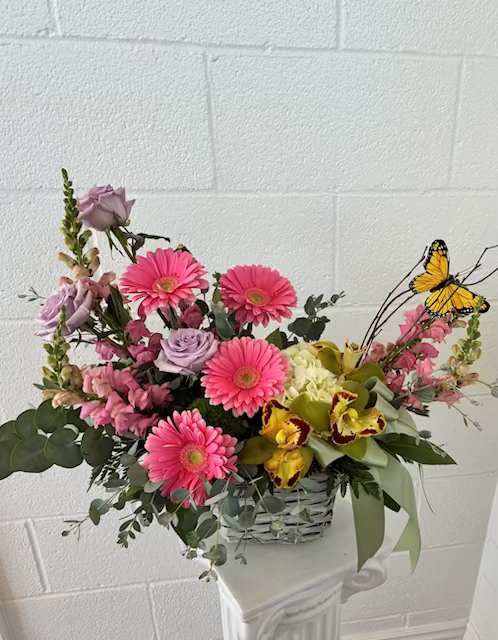 Flutter and Bloom  - A cheerful garden-style flower arrangement that reads like spring in a basket - perfect as a birthday bouquet, thank-you gift, or just-because surprise. This flower arrangement pairs bright pink gerbera daisies with soft lavender roses and yellow-green orchids to create a feminine, uplifting color story; decorative butterfly picks add a playful, whimsical touch that makes the recipient feel noticed and celebrated.  Arranged low in a woven basket, the bouquet has a full, outward-reaching shape thanks to smaller pink filler blooms and airy branching accents that add texture and movement. Colors: pink, purple, yellow, white; flowers include gerbera daisies, roses, orchids, and other seasonal fillers. A coordinating ribbon finishes the basket for easy display.  Ideal for celebrating a friend, mother, or colleague who loves bright, happy florals - the kind of arrangement that brings instant warmth to a kitchen table or office desk. No special care is needed beyond fresh water; to keep blooms looking their best, place in a cool spot away from direct sun and change the water every couple of days.