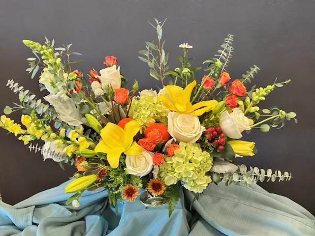 Nova Bloom - Bright yellow lilies take center stage in this airy garden-style flower arrangement, a cheerful birthday bouquet or congratulations gift that will make the person you're thinking of feel celebrated. The mix of white roses and warm orange spray roses against pale green hydrangeas creates a high-contrast, uplifting palette that reads sunny and festive - perfect for birthdays, new jobs, or simply saying "you're on my mind."  Tall branching stems and eucalyptus-like foliage give the design a loose, expansive silhouette with lots of movement and texture, while a clear glass vase lets the blooms float above clean, visible stems for a fresh, modern look. This large arrangement is ideal for someone who loves a natural, just-picked-from-the-garden feel; it arrives ready to display and brings bright color and gentle fragrance to any room.