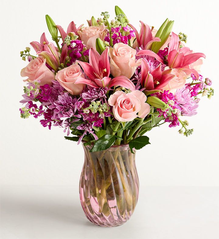 Mom™ Bouquet - Soft blush roses and open pink lilies create a romantic, feminine focal point in this Mom™ bouquet-perfect as a Mother's Day arrangement, birthday bouquet, or anniversary flowers to make her feel truly cherished. Purple chrysanthemums and bright magenta filler blooms add lively contrast and depth, so the person you're celebrating will feel seen and celebrated the moment the vase arrives.  This lush, rounded bouquet is densely layered for a full, polished look, with upright lily buds adding height above the softer rose heads and clustered accent flowers woven throughout. Arranged in a ribbed pink-tinted glass vase, the combination of pink, purple, white, and green tones reads fresh and elegant-an ideal choice for someone who loves soft pastels with a pop of color.  Designed for gifting, it's the kind of bouquet that marks special moments-Mother's Day, birthdays, or anniversaries-while brightening a kitchen table or bedside. The sturdy glass vase makes setup easy; trim stems and refresh the water every few days to keep blooms looking their best.