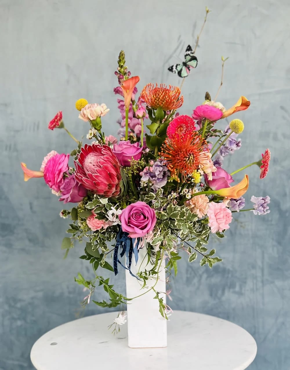Georgiana - A bright, contemporary floral arrangement designed in a tall vase, featuring vibrant shades of pink, orange, and lavender. This modern composition blends bold color with elegant structure, creating height, movement, and visual impact.  Each arrangement is handcrafted with premium seasonal blooms, carefully selected for their tone, texture, and natural form. The vertical design enhances the sense of flow, allowing each flower to stand out while working together in a cohesive, expressive arrangement.  Flowers and greenery may vary due to seasonal availability, but that’s part of the charm! Our creative designers will substitute with the freshest high end product available- some grown in our very own garden!- and create something beautiful.  *Have a special request?* Let us know in the special instructions section during checkout.
