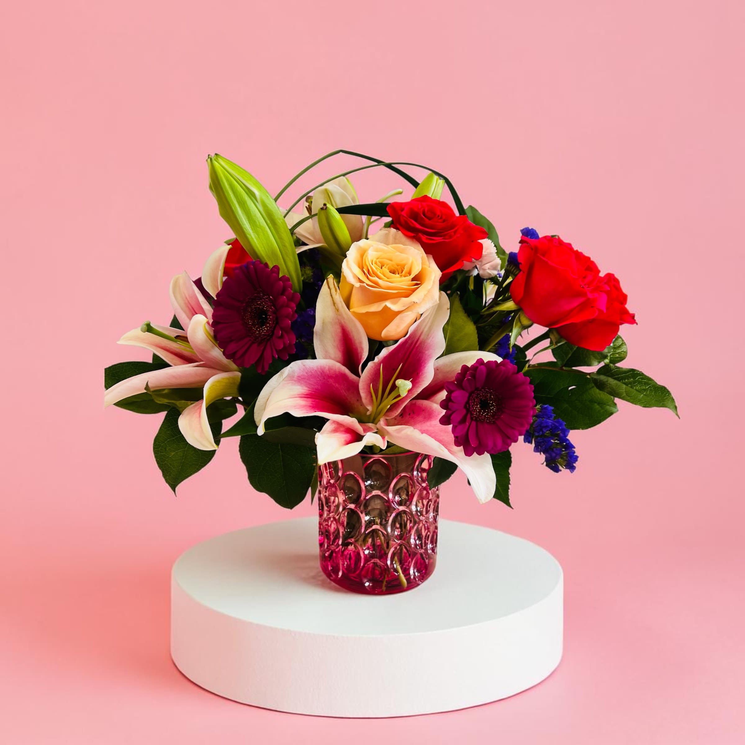 Mom Special - Pink, red, white and purple roses, lilies and gerbera in a rounded, gift-ready bouquet with greenery, accented by blue blooms and a decorative pink glass vase - warm, lively, and affectionate.
