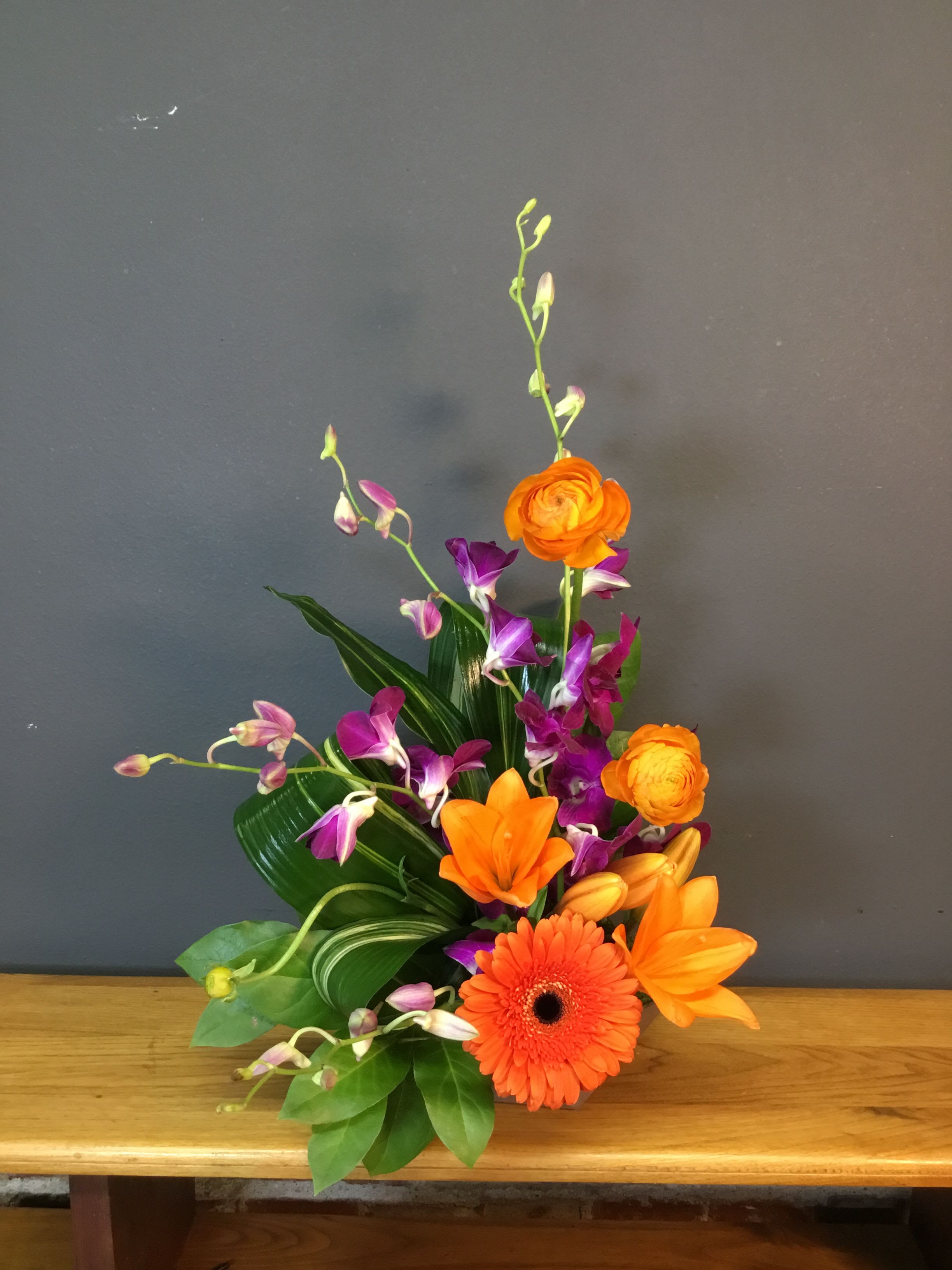 Tropical Dream - Bright, celebratory color pops make this Tropical Dream flower arrangement a joyful choice for a birthday bouquet, Mother's Day gift, or a bold "thinking of you" surprise. The mix of bright orange gerbera daisies and lily-like blooms with vivid purple and pink orchid sprays gives the arrangement an instantly tropical, modern feel that reads as festive for anniversaries, housewarmings, or a cheerful "just because" moment.  This design layers a large orange gerbera at the front with round ranunculus-style blooms and taller orange blossoms to create height and texture, while glossy green leaves form a lush base that makes the colors sing. Purple and pink orchids arc through the composition for contrast and an elegant touch, producing a lively, energetic silhouette ideal for congratulating someone, saying thanks, or offering comfort during sympathy and funeral remembrances with tasteful color.  Made to stand out on a mantel, entry table, or office desk, Tropical Dream suits seasonal spring celebrations as well as year-round occasions like Father's Day, weddings, or "I'm sorry" gestures. Care is simple: keep water fresh and trim stems as needed to preserve the vivid colors and structure so the recipient feels genuinely celebrated every time they see it.