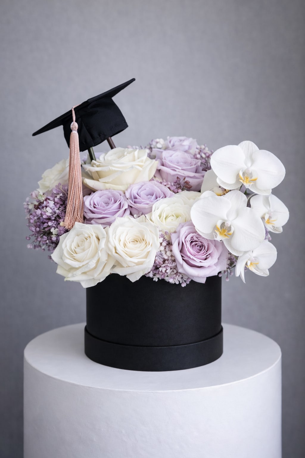 Lavender Graduation Luxe – Black Box - A sophisticated floral arrangement featuring premium white and lavender roses paired with elegant white orchids, designed in a modern black keepsake box for a bold and elevated look. Finished with a graduation cap detail, this design offers a striking contrast that feels both contemporary and timeless.  Perfect for those who love a clean, high-end aesthetic with a celebratory touch.  Bouquet will be delivered approximately as shown in the picture. Flower varieties and shades may vary based on availability while maintaining the overall style, color palette, and quality.  Same-day flower delivery available in Southwest Florida, including Naples, Bonita Springs, and Fort Myers Beach.  Best for:Graduation gifts