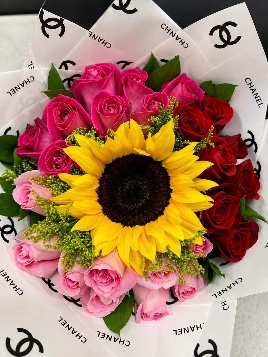 Sunshine Love  - A single, bold sunflower anchored amid a tight cluster of pink and red roses makes this celebratory bouquet a joyful mix of sunshine and romance-perfect as a birthday bouquet, anniversary surprise, or "thinking of you" gift. The bright yellow center draws the eye instantly so the person you're sending it to will feel noticed and celebrated the moment the wrapping is opened.  This full, rounded arrangement pairs velvety red and soft pink roses around the dominant sunflower for striking contrast, with the colors balanced by the crisp white decorative wrapping paper printed with a chic black logo. Finished with a decorative paper wrap, the presentation reads gift-ready and polished without extra fuss.  Give this to someone who loves vibrant, fashion-forward florals-she'll feel truly seen and cheered. Bouquet size is a generous hand-tied arrangement; to keep blooms fresh, trim stems and change the water every other day, and place away from direct sunlight and heat sources.