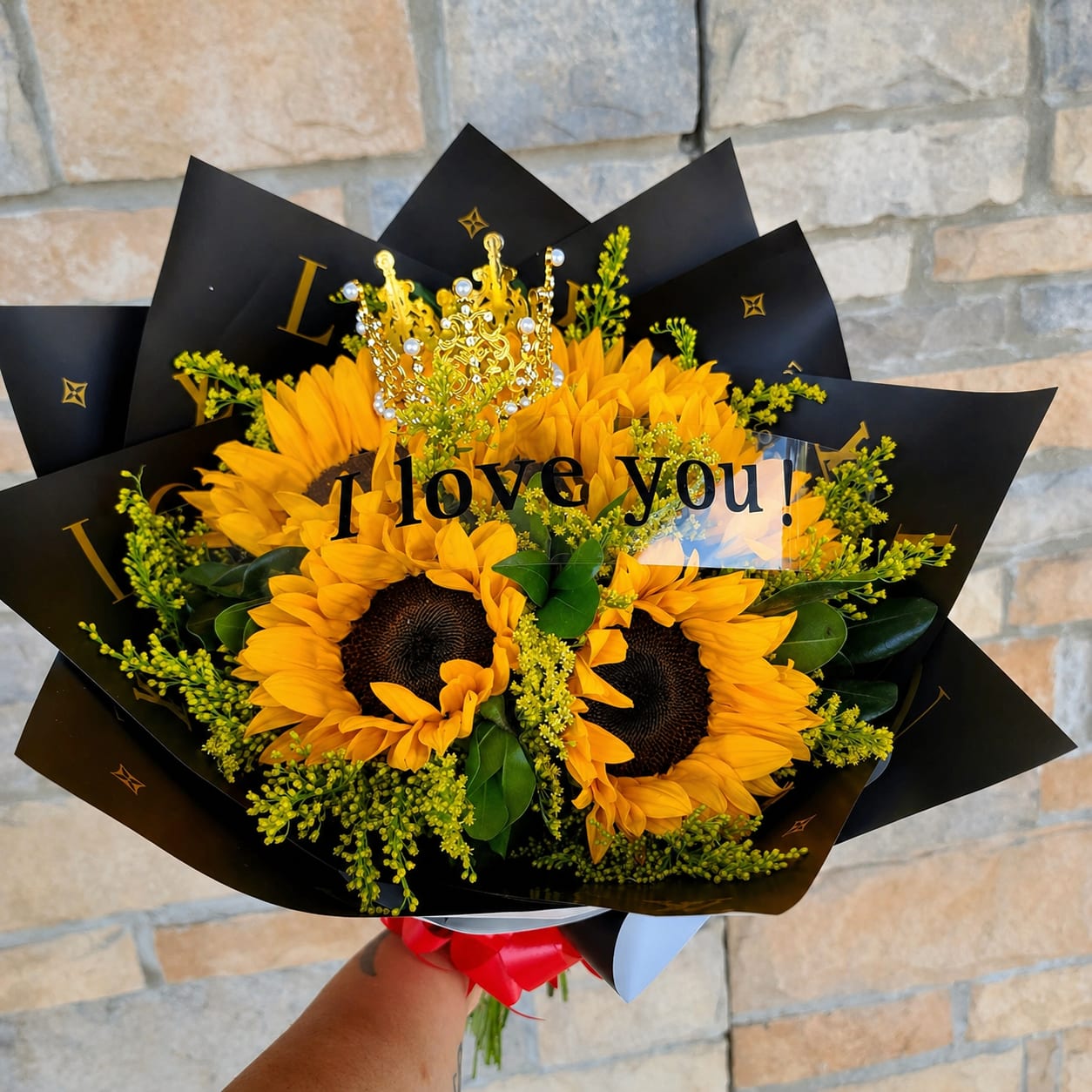  Golden Crown  - A cluster of golden sunflowers takes center stage in this bold love-themed bouquet - a cheerful anniversary or "just because" flower arrangement that reads as both romantic and celebratory. The bright yellow petals and dark brown centers pop against matte black floral wrap printed with gold lettering and star motifs, while a decorative crown topper and a printed "I love you!" sign make the gift message unmistakable.  Tightly arranged sunflowers are complemented by small yellow filler blooms and glossy green foliage for texture and fullness. Finished with a red ribbon and a jeweled crown pick, the presentation feels like a keepsake moment - vibrant, confident, and joyful. Ideal for the person you want to make feel truly seen on birthdays, anniversaries, or spontaneous romantic gestures; arrives wrapped and ready to present. Care: keep stems in fresh water, trim stems at an angle, and refresh water every other day to extend vase life.