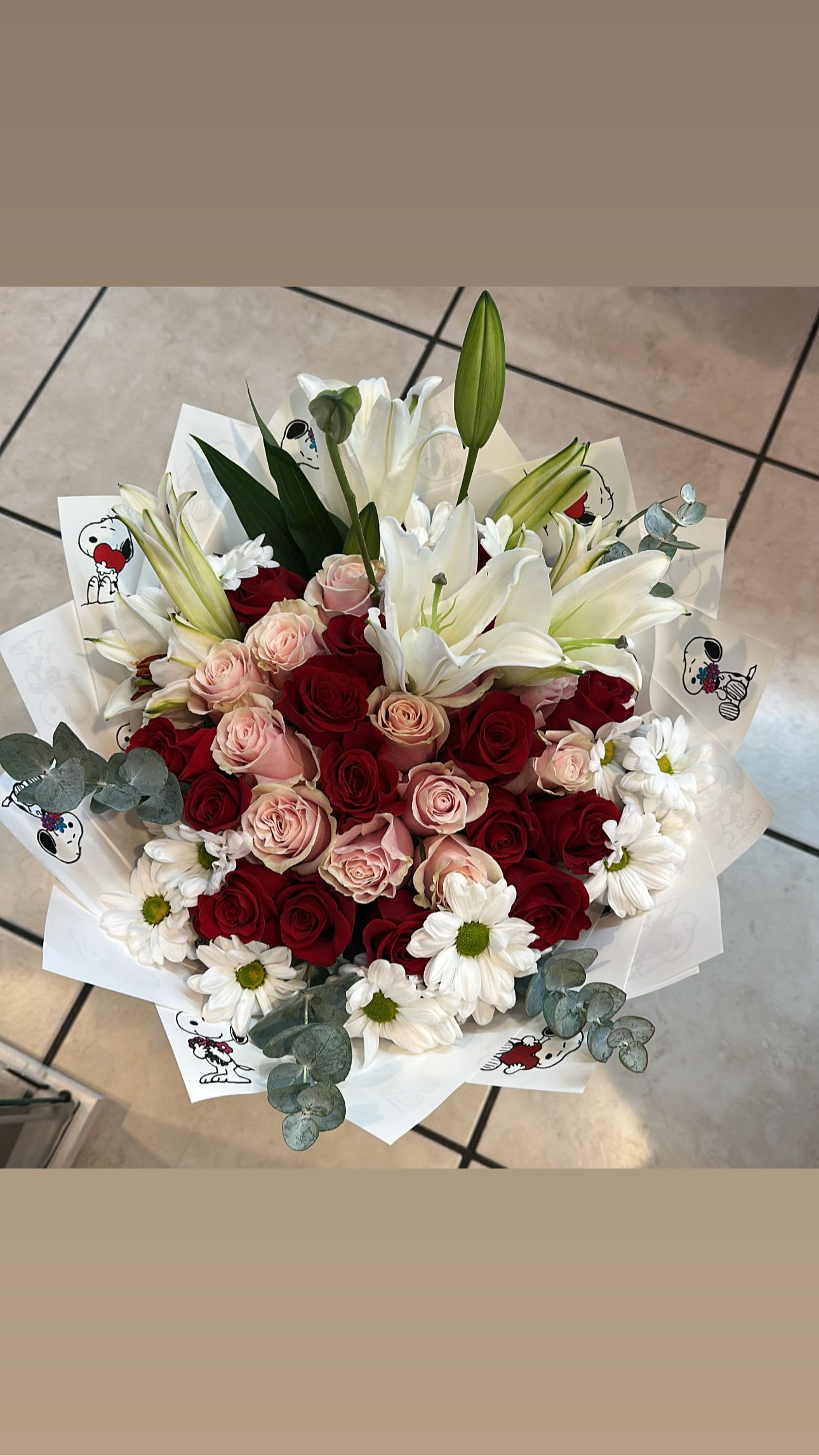 Snoppy fest  - A lush, rounded bouquet that pairs dense red and soft blush roses with large white lilies and scattered white daisies - a romantic flower arrangement perfect for anniversaries, Valentine's Day, or any special-occasion gifting. The lilies rise above the rose cluster to create graceful height and contrast, so the person you're thinking of will feel seen and celebrated the moment the bouquet is set before them.  This arrangement features red, pink and white blooms accented by pale eucalyptus-like greenery for a fresh, airy texture. It arrives wrapped in white printed decorative paper for a gift-ready presentation that reads both elegant and joyful. Sized to make an impression without overwhelming a table, it's an ideal choice when you want to say "I love you," "congratulations," or "I'm thinking of you" with classic floral romance.