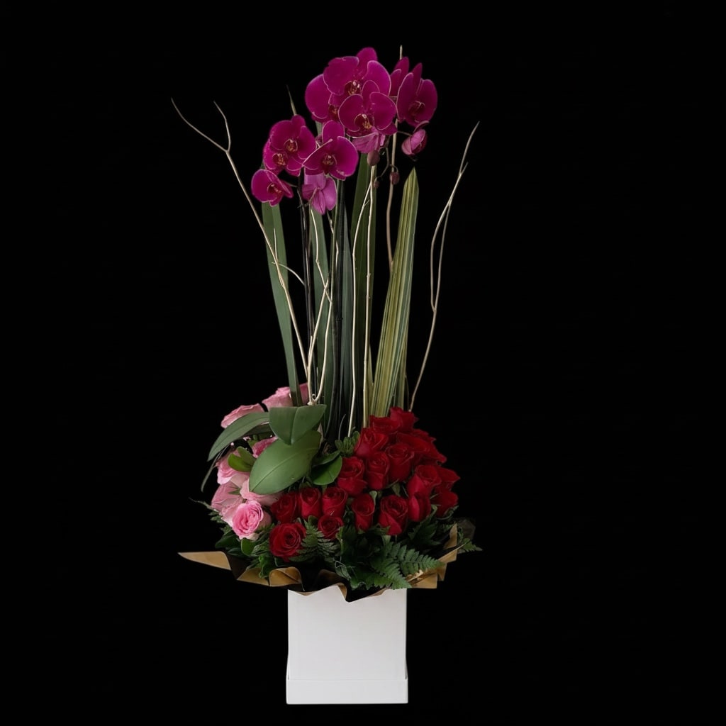 You are everything in the world - A vertical, romantically bold arrangement that reads like a declaration: dense red roses form a lush base while pink roses and magenta Phalaenopsis orchids soar above, creating a dramatic birthday bouquet or anniversary arrangement that makes the person you're thinking of feel truly adored. The tall, reed-like accents and broad glossy leaves add architectural height and movement, so she'll notice it from across the room and feel celebrated the moment it arrives.  Arranged in a crisp white presentation box with decorative wrap, the composition balances intense reds and soft pinks with touches of purple and white for depth and contrast. The compact base of roses gives rich texture and scent, while the orchids provide elegant vertical lines; overall dimensions present as a tall, eye-catching centerpiece perfect for intimate dinners or formal celebrations. Designed as a premium gift, it arrives ready-to-display - simply place on a stable surface away from direct heat and change the water source if provided to keep blooms fresh longer.