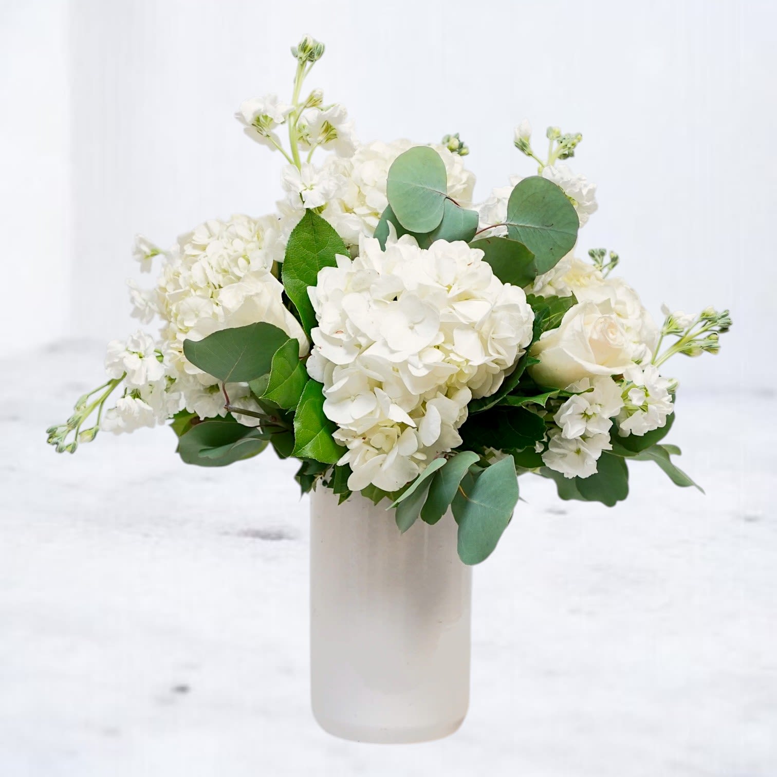 Milkglass - Milkglass is a modern white flower arrangement designed with lush white hydrangea, premium roses, delicate stock, and fresh eucalyptus in a soft garden-style composition. Designed in a sleek ceramic vase, this arrangement blends layered white florals with fresh greenery for a clean, timeless aesthetic filled with texture and natural movement.  Elegant yet understated, Milkglass is perfect for sympathy flowers, celebrations of life, birthdays, thank you gifts, new baby gifts, anniversaries, or everyday flower delivery. Its airy monochromatic palette and modern editorial styling make it a versatile design suited for both contemporary and classic spaces.  Handcrafted with premium seasonal flowers and available for same-day flower delivery in Magnolia, TX and surrounding areas including The Woodlands, Montgomery, Conroe, Spring, Tomball, and nearby Lake Conroe communities.