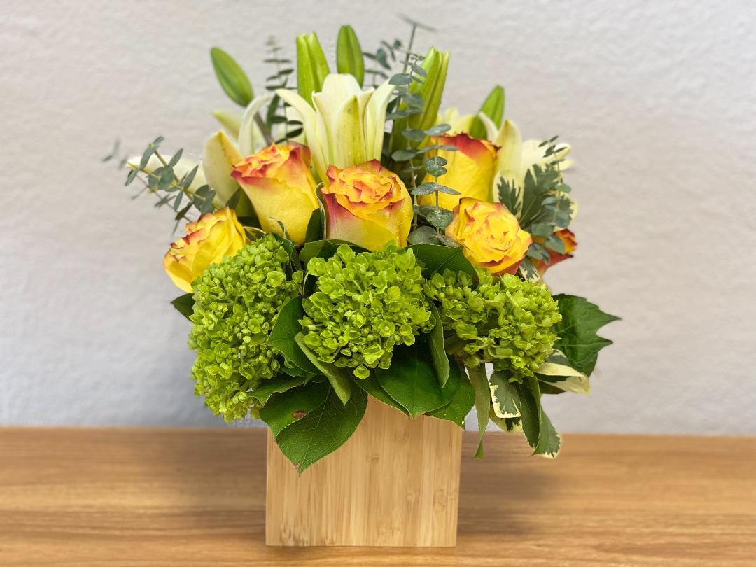 Yellow roses and white lilies in a wooden cube vase with green hydrangeas