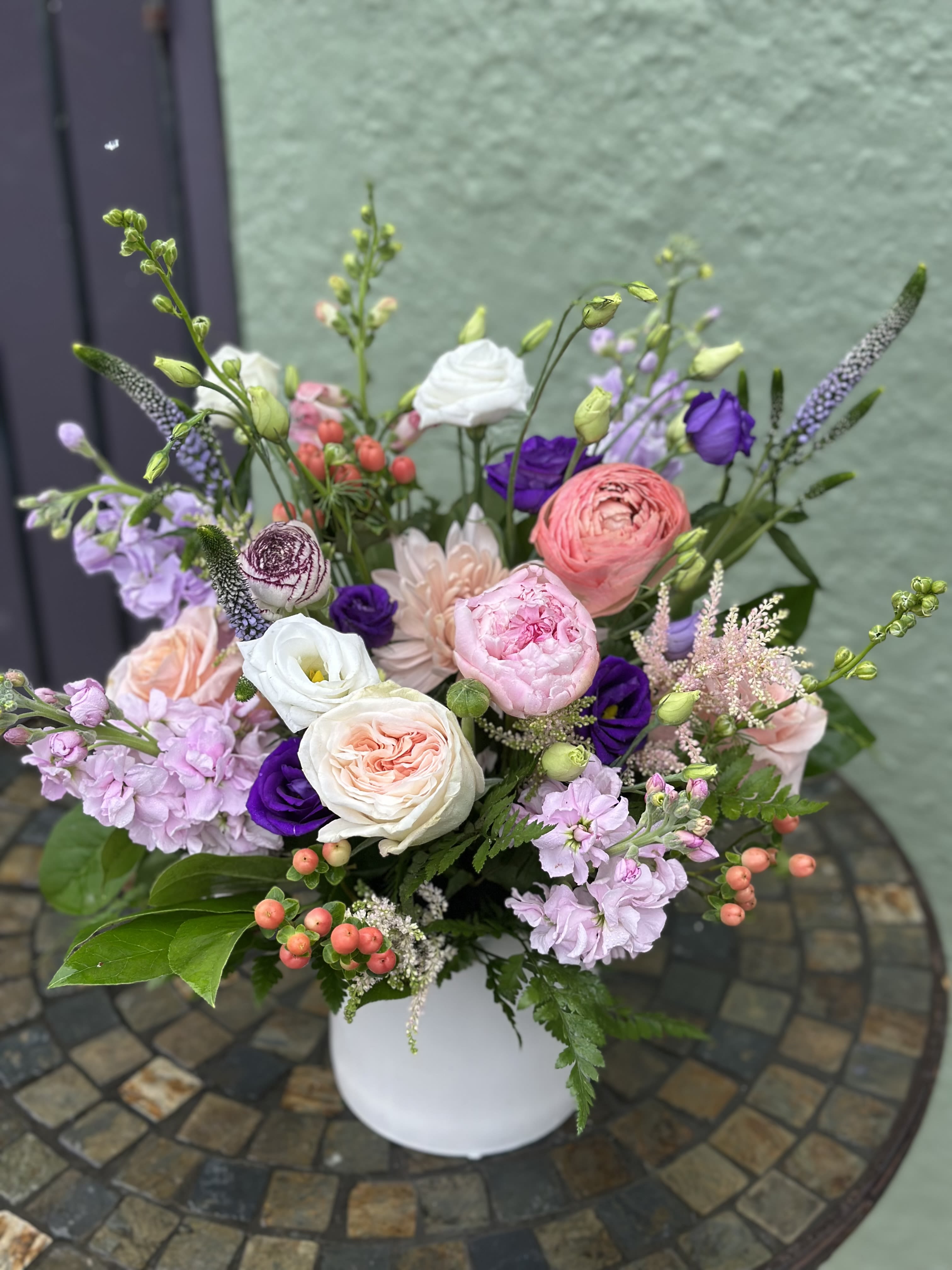 Pastel pink and white flower arrangement with purple accents in a white vase on a mosaic table.