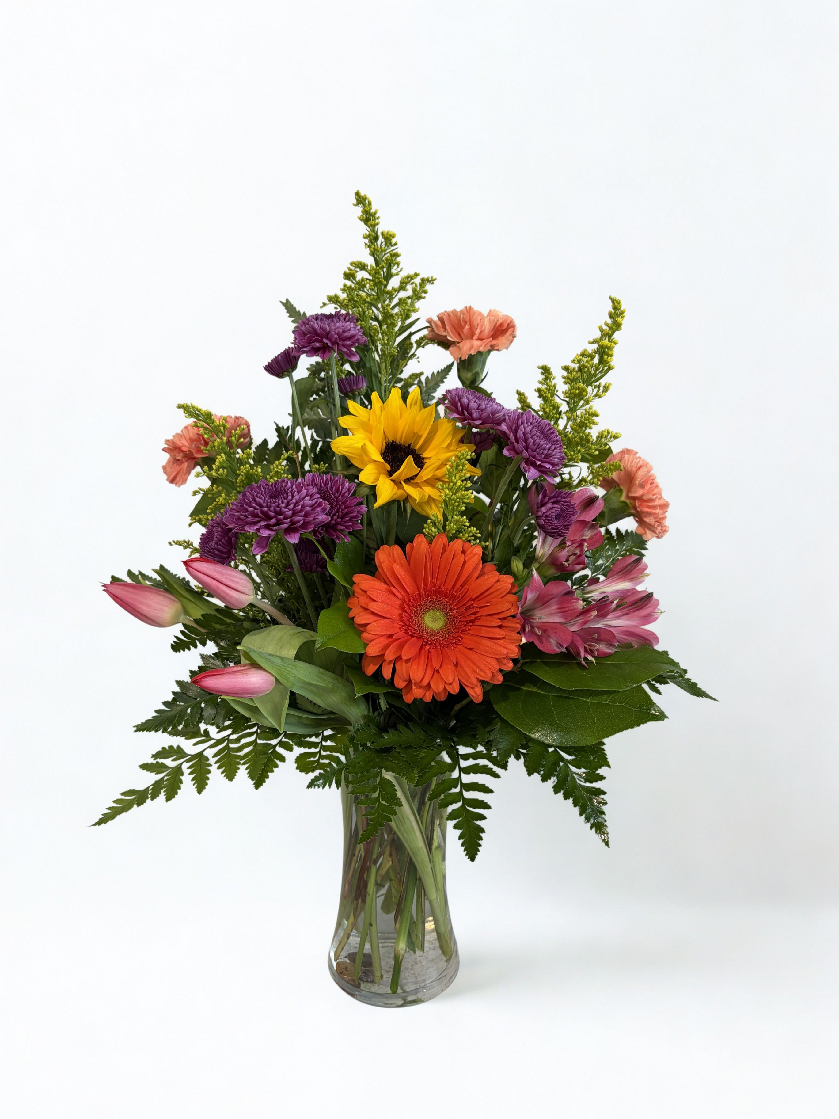 Sunset Chaser - A lively garden-style bouquet that brightens any birthday, Mother's Day, or "just because" moment - Sunset Chaser pairs sunny sunflowers and orange gerbera daisies with soft pinks, blues, and lavenders for an uplifting mixed bouquet. Designed to make the person you're thinking of feel celebrated, the arrangement reads cheerful and gentle at once: bold yellow focal blooms catch the eye while cool blues and purples soothe the palette.  Packed into a clear glass vase, this upright arrangement layers rounded hydrangea, sunflowers, gerbera, lilies, snapdragons, alstroemeria, carnations, and chrysanthemums with airy yellow filler and fern-like greenery for texture and movement. The size is full and garden-like, with taller spikes adding height and rounded blooms creating a lush center - an ideal pick for spring, thank-you gifts, congratulations, or any day you want someone to smile.
