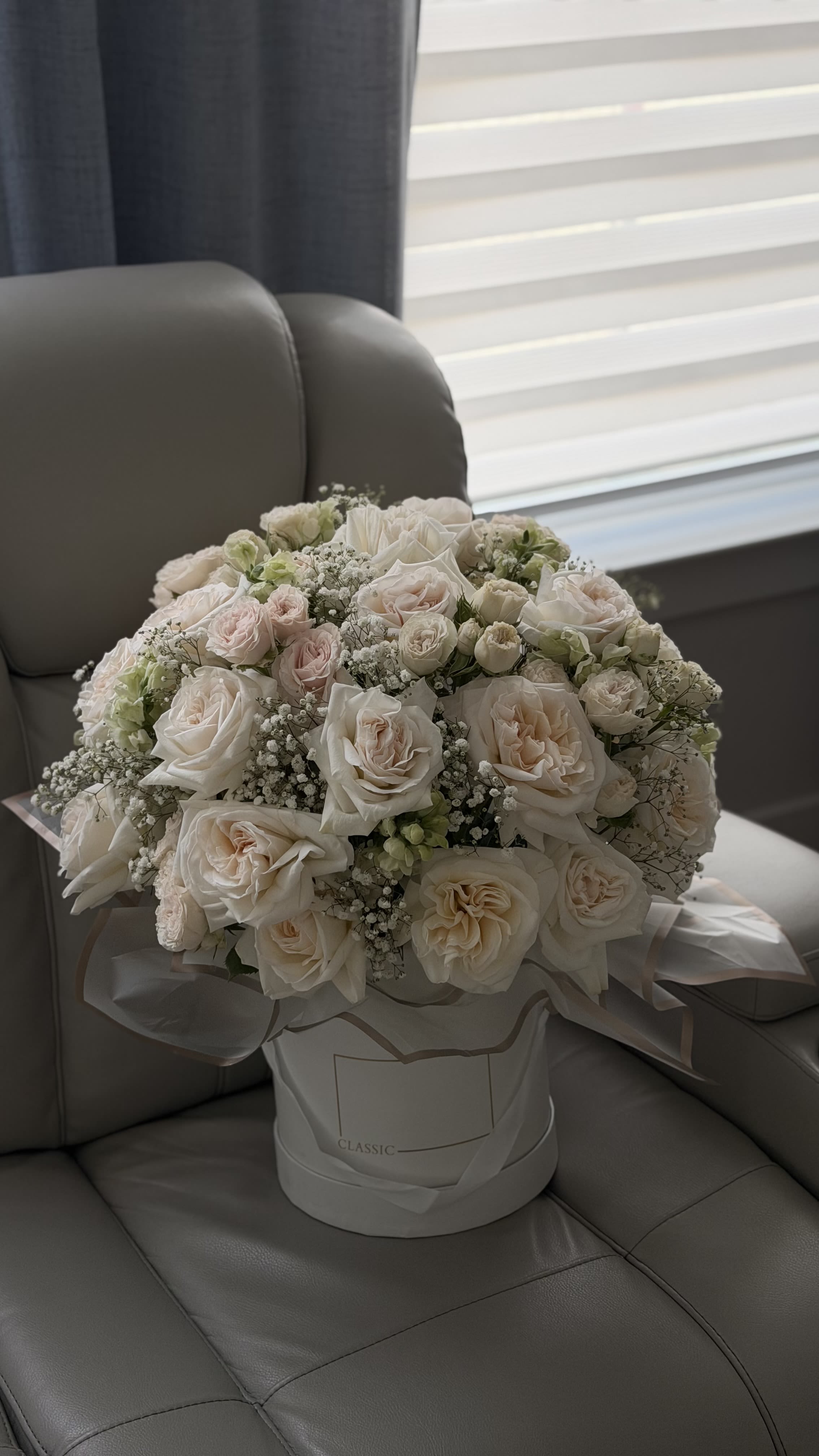 Ivory Whisper - A timeless expression of elegance and quiet luxury. Ivory Whisper is composed of premium garden roses and spray roses in soft ivory and blush tones, delicately accented with airy baby’s breath for a light, ethereal finish.  Designed in a refined hat box, this arrangement feels polished and effortlessly sophisticated. The soft layering of petals creates a full, cloud-like silhouette while maintaining a clean, graceful look.